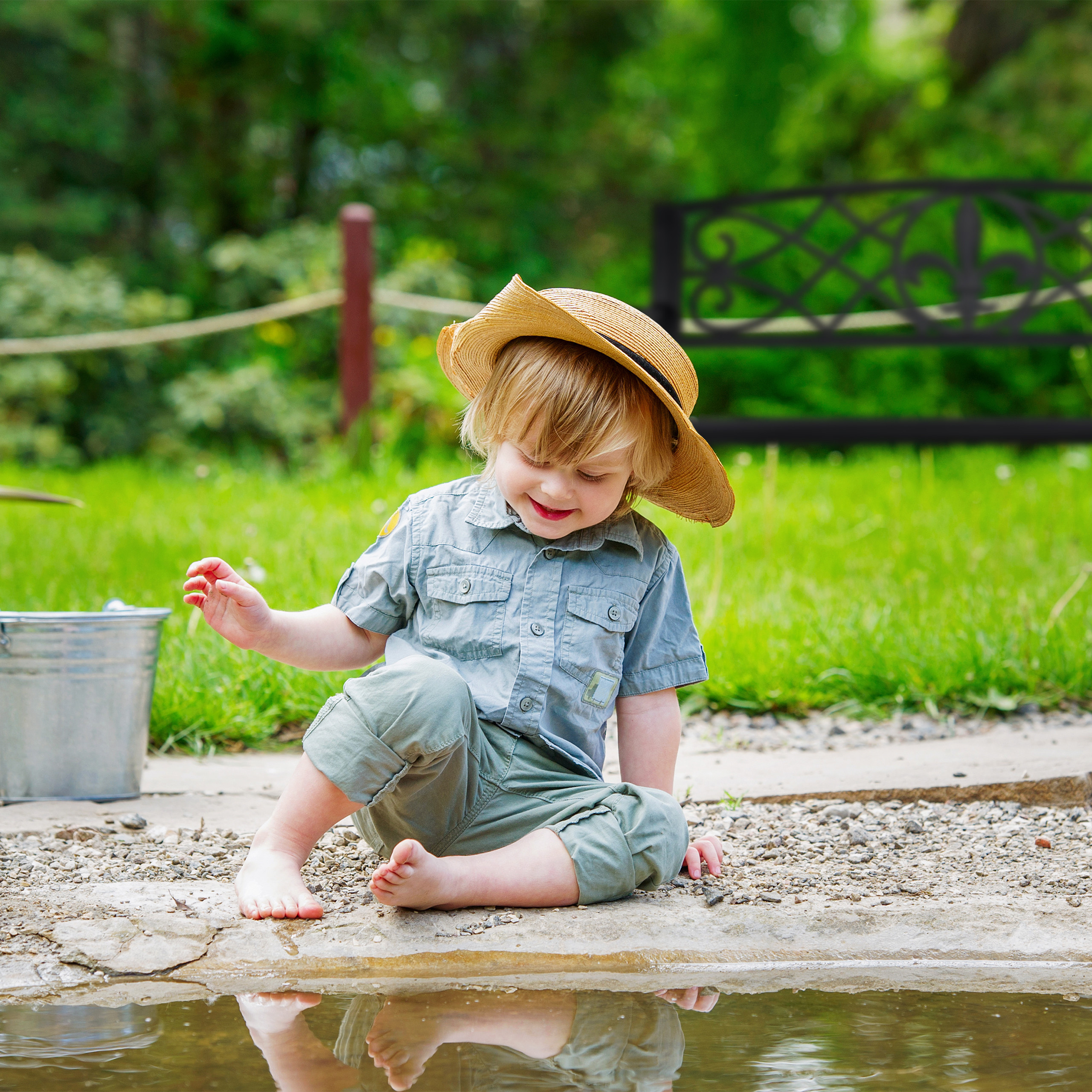 Kleiner Junge mit Strohhut sitzt am Teich und betrachtet sein Spiegelbild.