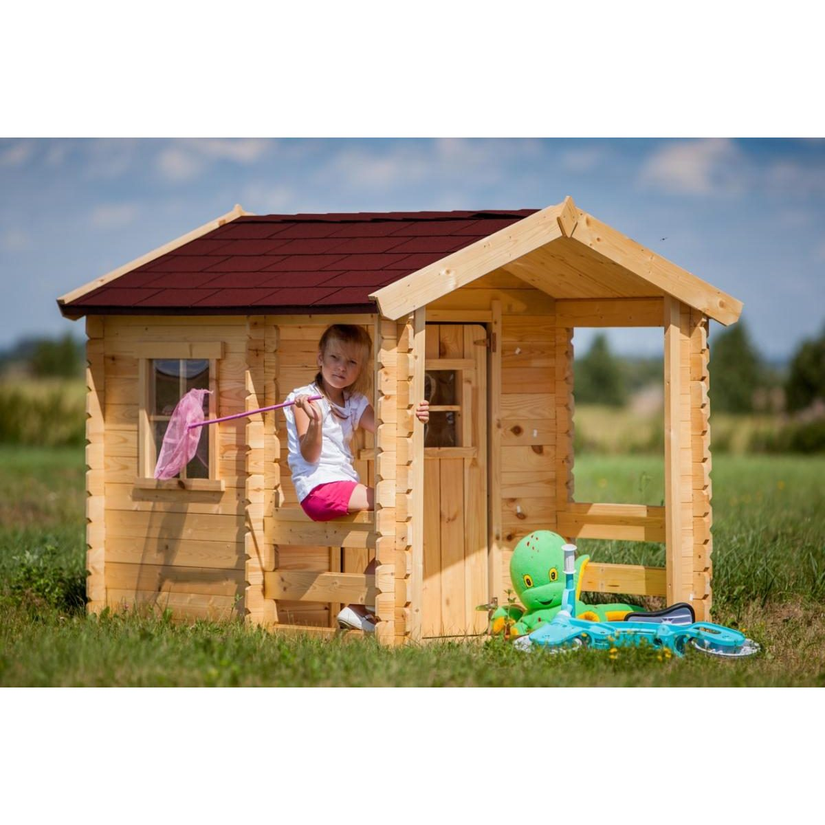 Ein kleines Mädchen sitzt in einem Holzspielhaus im Garten, hält einen rosa Schmetterlingskescher und ein grünes Kuscheltier.