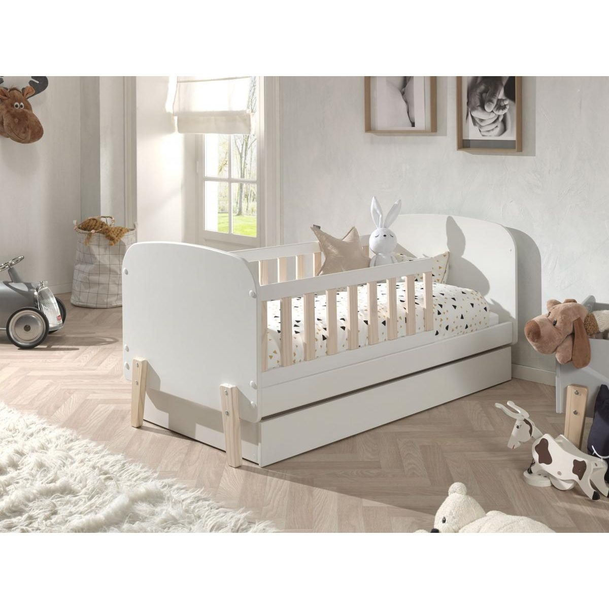 Ein weißes Kinderbett mit Holzbeinen und Gitterstäben in einem Kinderzimmer, umgeben von Spielzeug und einem Fenster.