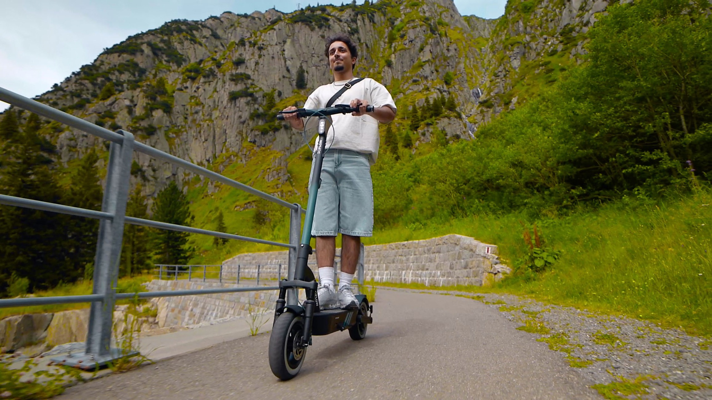 Ein lächelnder junger Mann fährt mit einem E-Scooter auf einem asphaltierten Weg vor einer Bergkulisse.