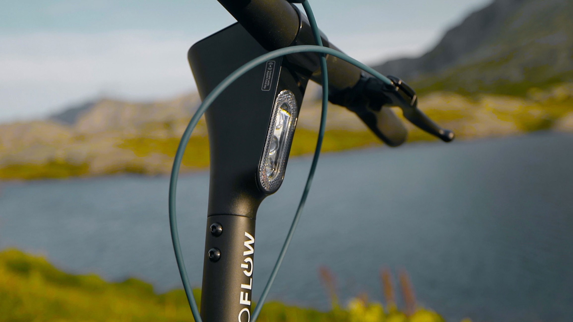 Nahaufnahme des Lenkers und der Vorderlampe eines schwarzen E-Scooters vor einer Berg- und Seenlandschaft.
