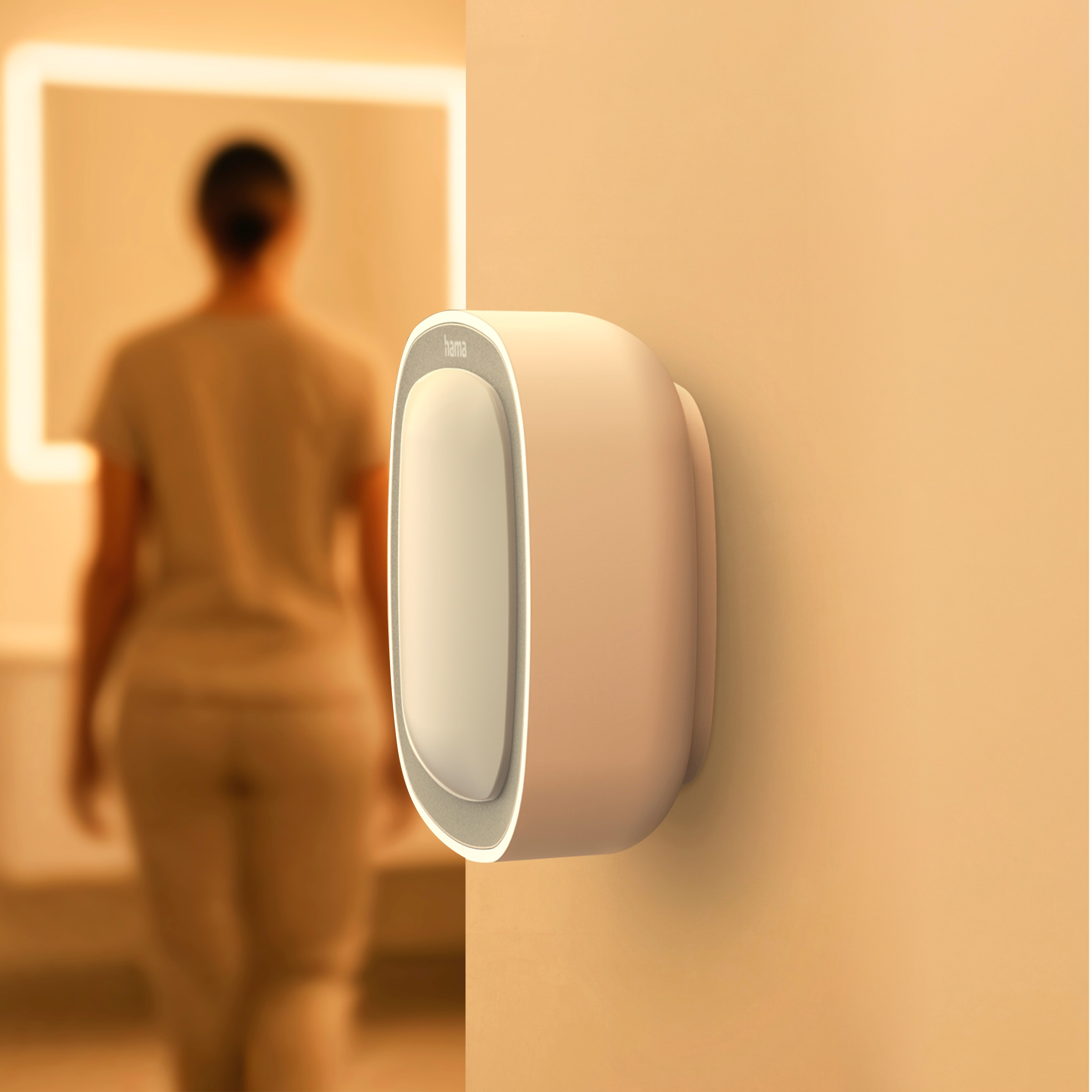 Ein weißes Hama-Smart-Gerät an einer Wand; im Hintergrund eine Person in einem warm beleuchteten Raum.