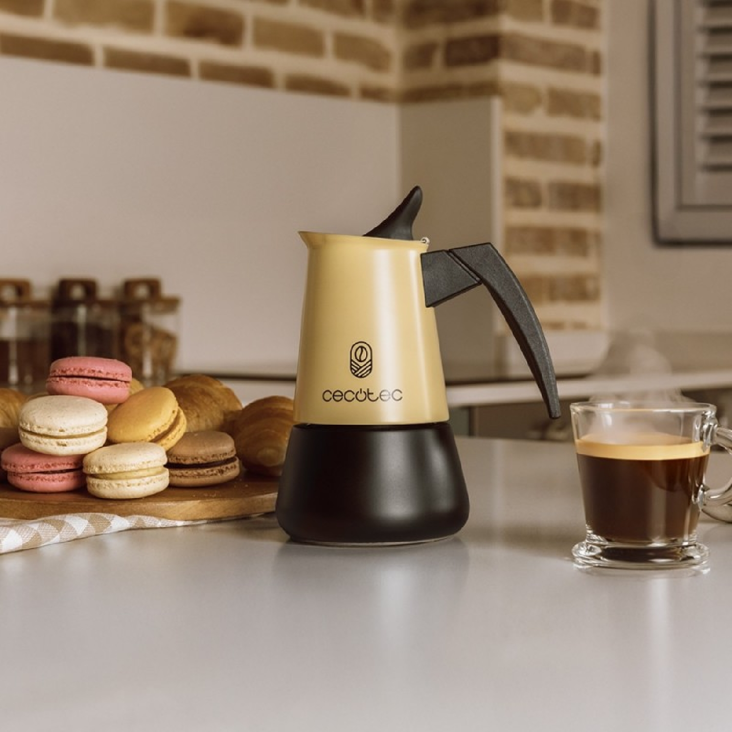 Una caffettiera Cecotec beige, una tazza di caffè e dei macaron su un bancone della cucina.