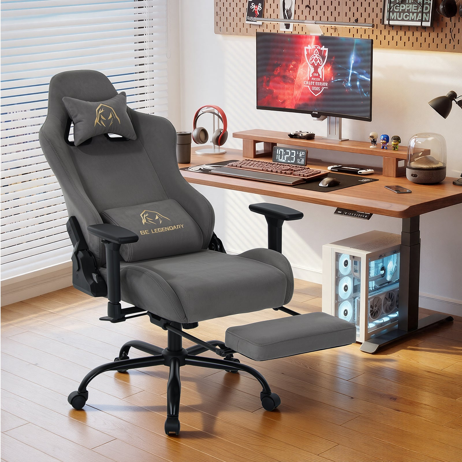 Ein grauer Gaming-Stuhl mit ausklappbarer Fußstütze steht vor einem Holztisch mit PC und Monitor in einem hellen Zimmer.