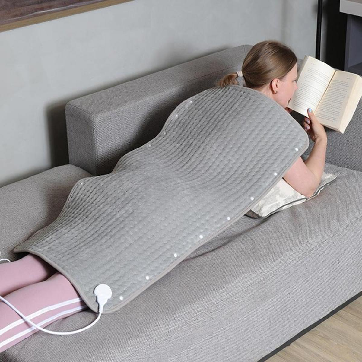 Eine Frau liegt auf einem grauen Sofa und liest ein Buch, während eine graue Heizdecke auf ihrem Rücken liegt.
