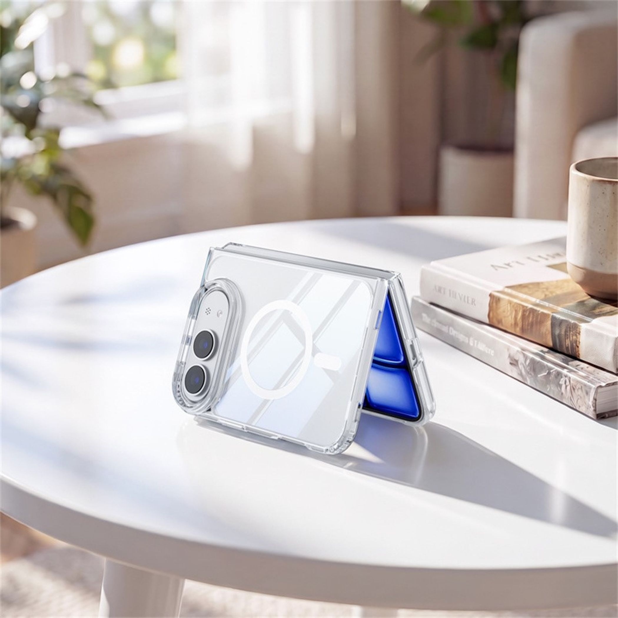 Ein faltbares Smartphone in einer klaren Hülle mit MagSafe-Ring steht auf einem weißen Tisch neben Büchern und einer Tasse.