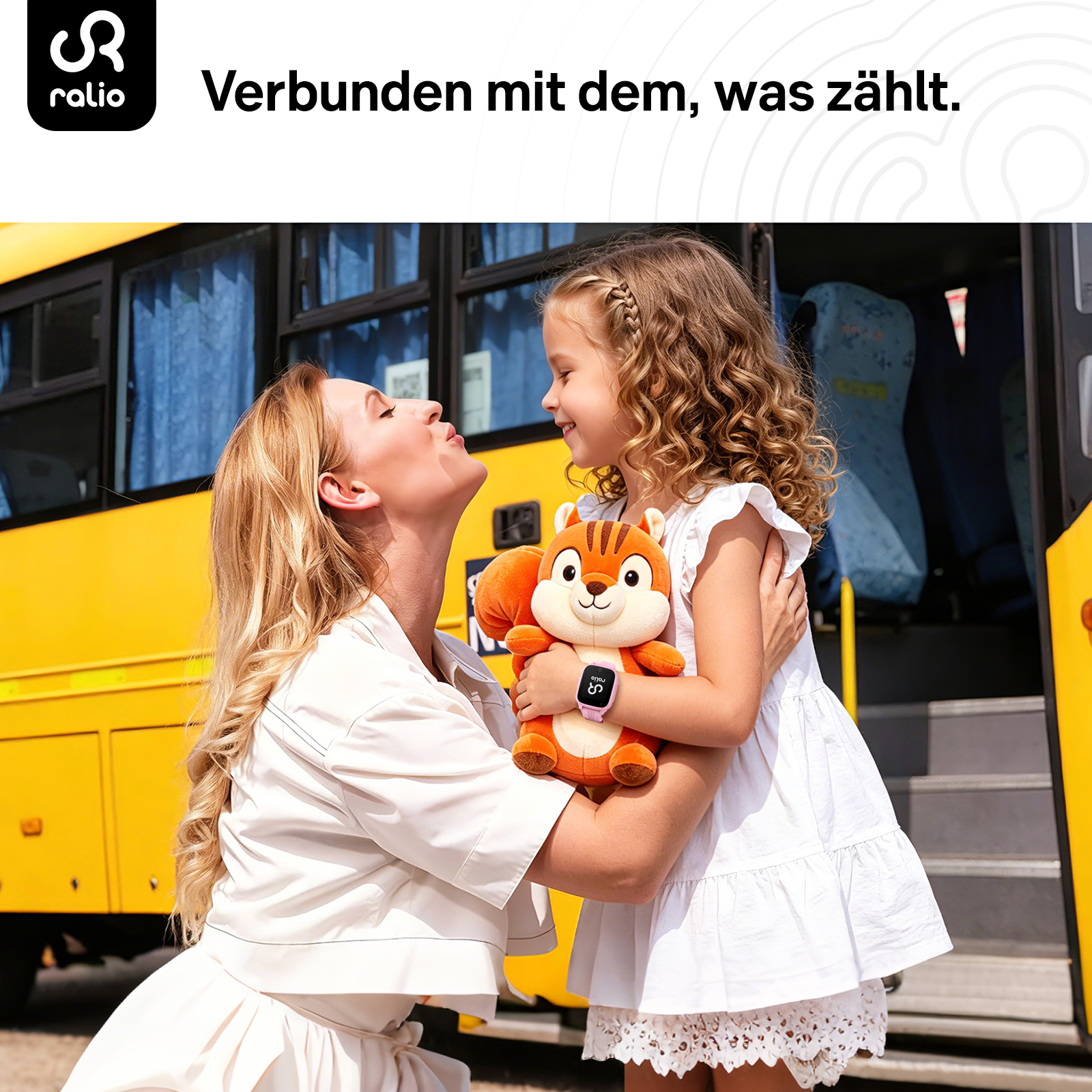 Mutter küsst ihre Tochter vor einem gelben Schulbus. Das Mädchen hält ein Plüschtier und eine Smartwatch.