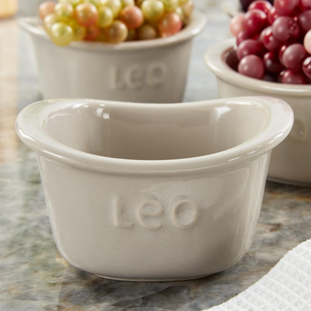 Trzy beżowe ceramiczne miski z napisem "Leo" na marmurze, dwie z nich wypełnione zielonymi i czerwonymi winogronami.