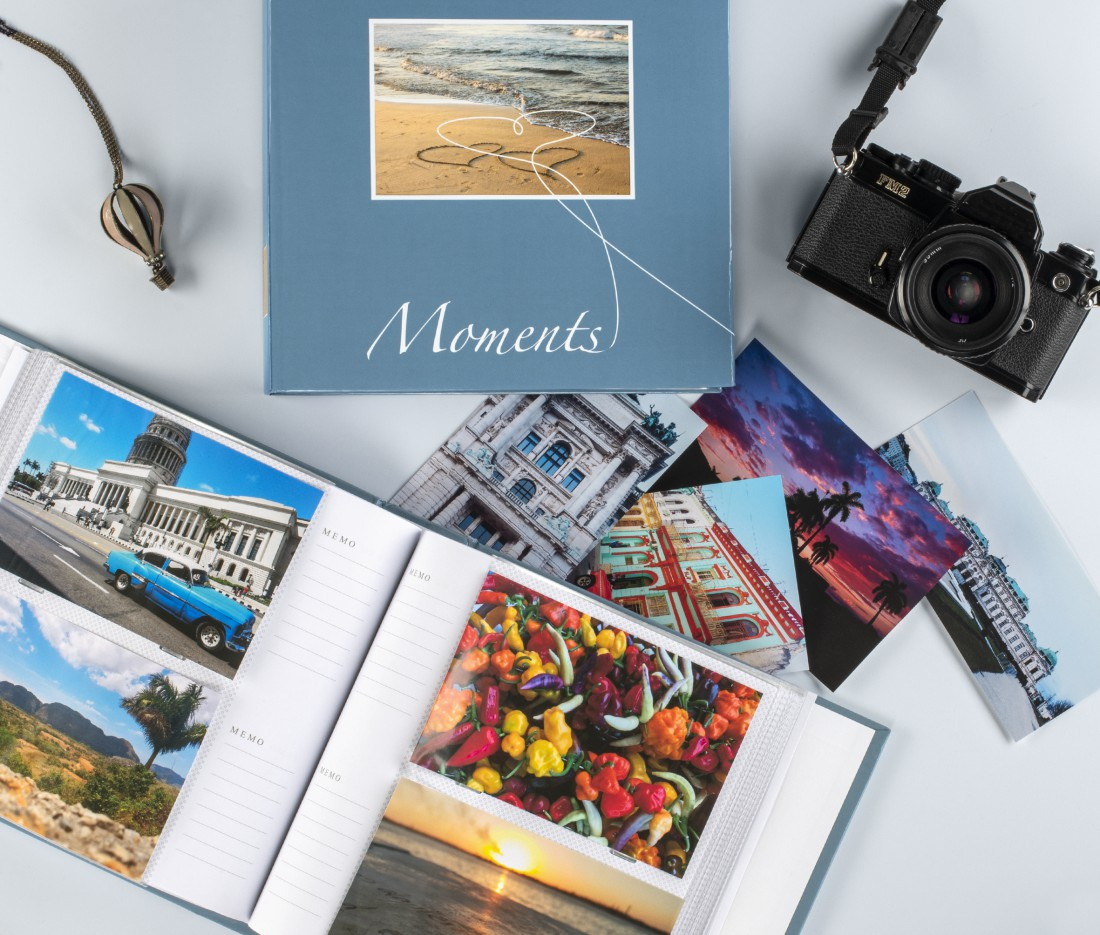 Blaue Fotobox mit Fotoalbum, Kamera, Anhänger und vielen bunten Reisefotos auf grauem Hintergrund.