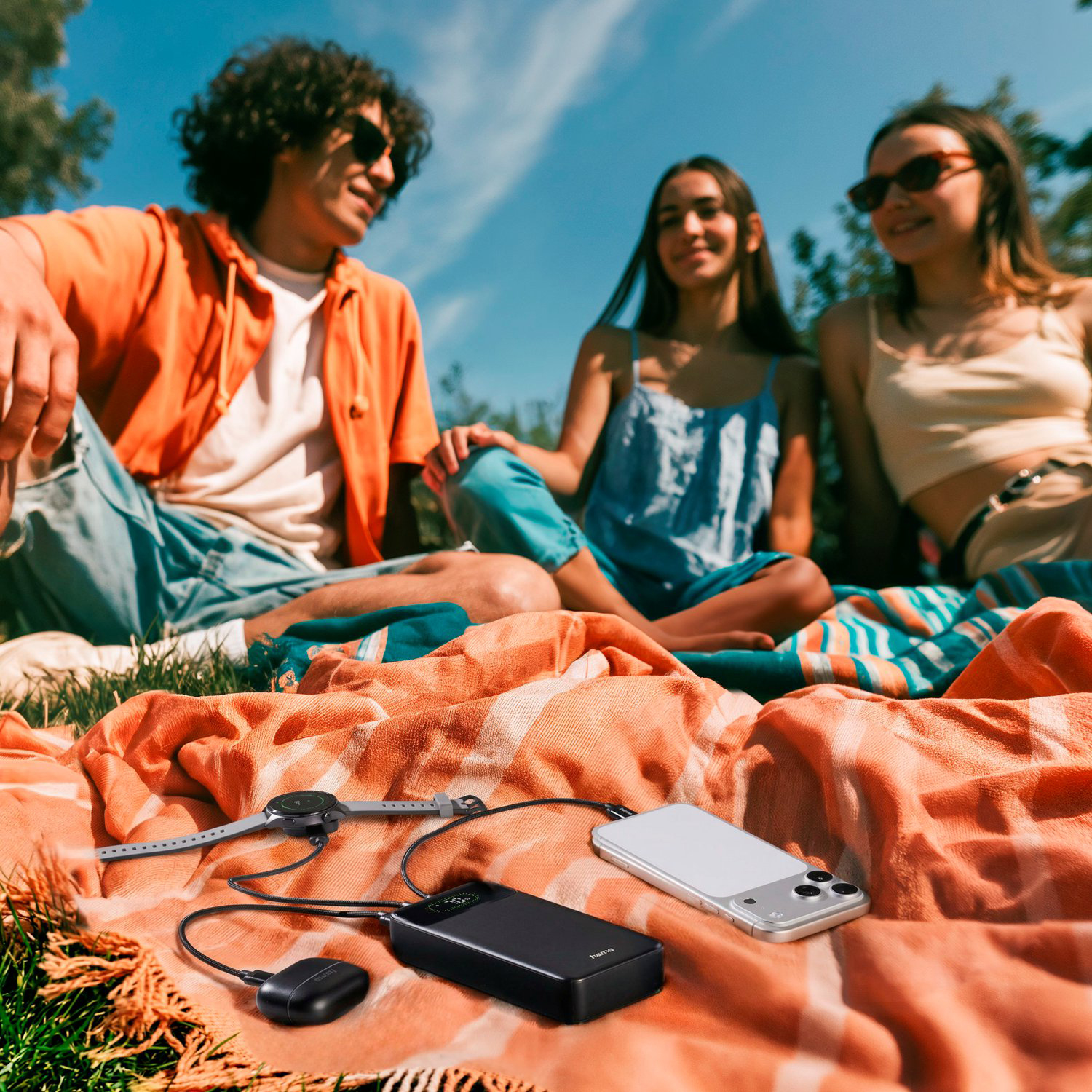 Drei Freunde entspannen im Freien, während eine Powerbank ein Telefon, eine Uhr und Ohrhörer lädt.