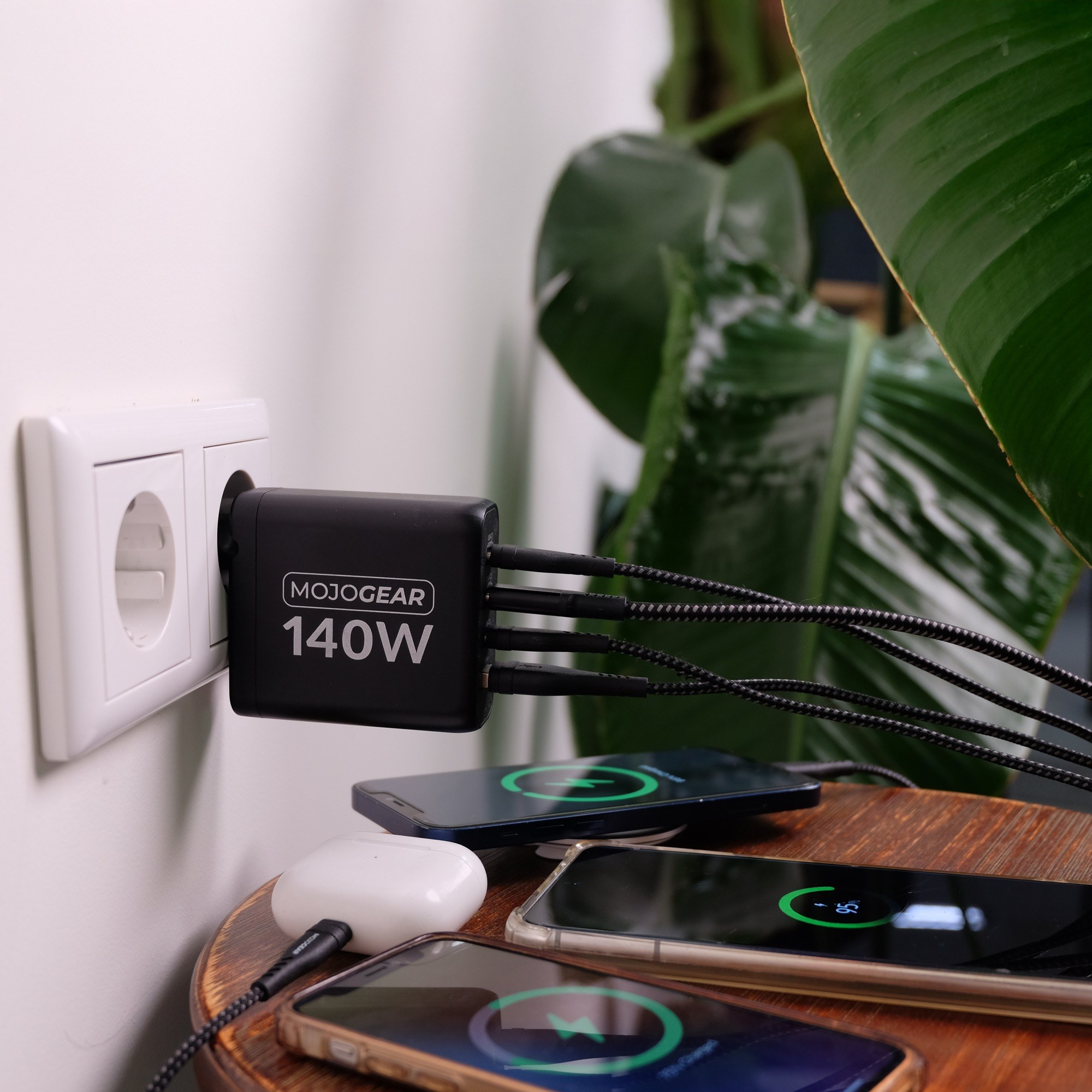 A black 140W Mojogear wall charger plugged into an outlet charges three smartphones and wireless earbuds on a wooden table.