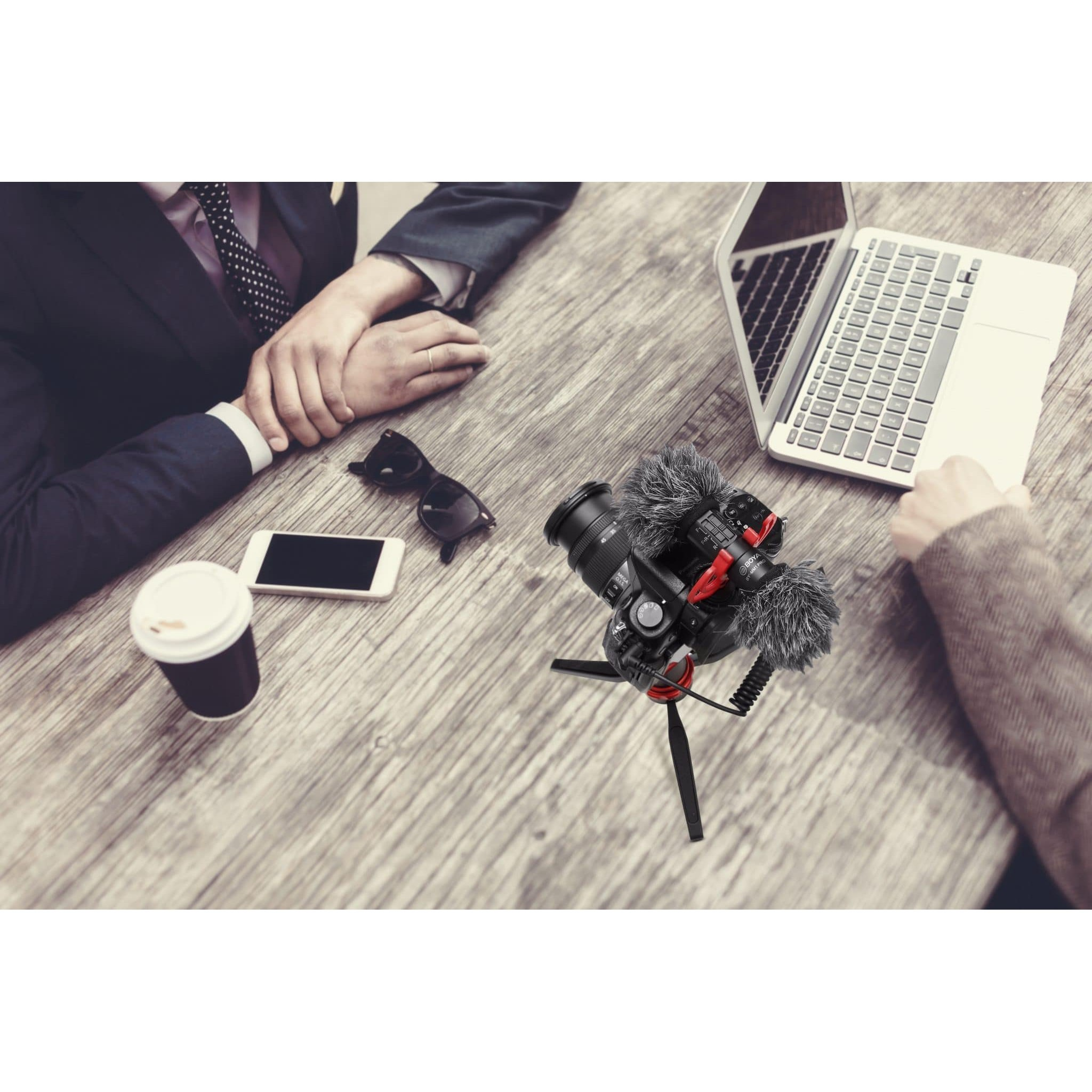 Ein Schreibtisch mit Kamera auf Stativ, Laptop, Smartphone, Sonnenbrille und Kaffeetasse, zwei Personen sitzen nah.