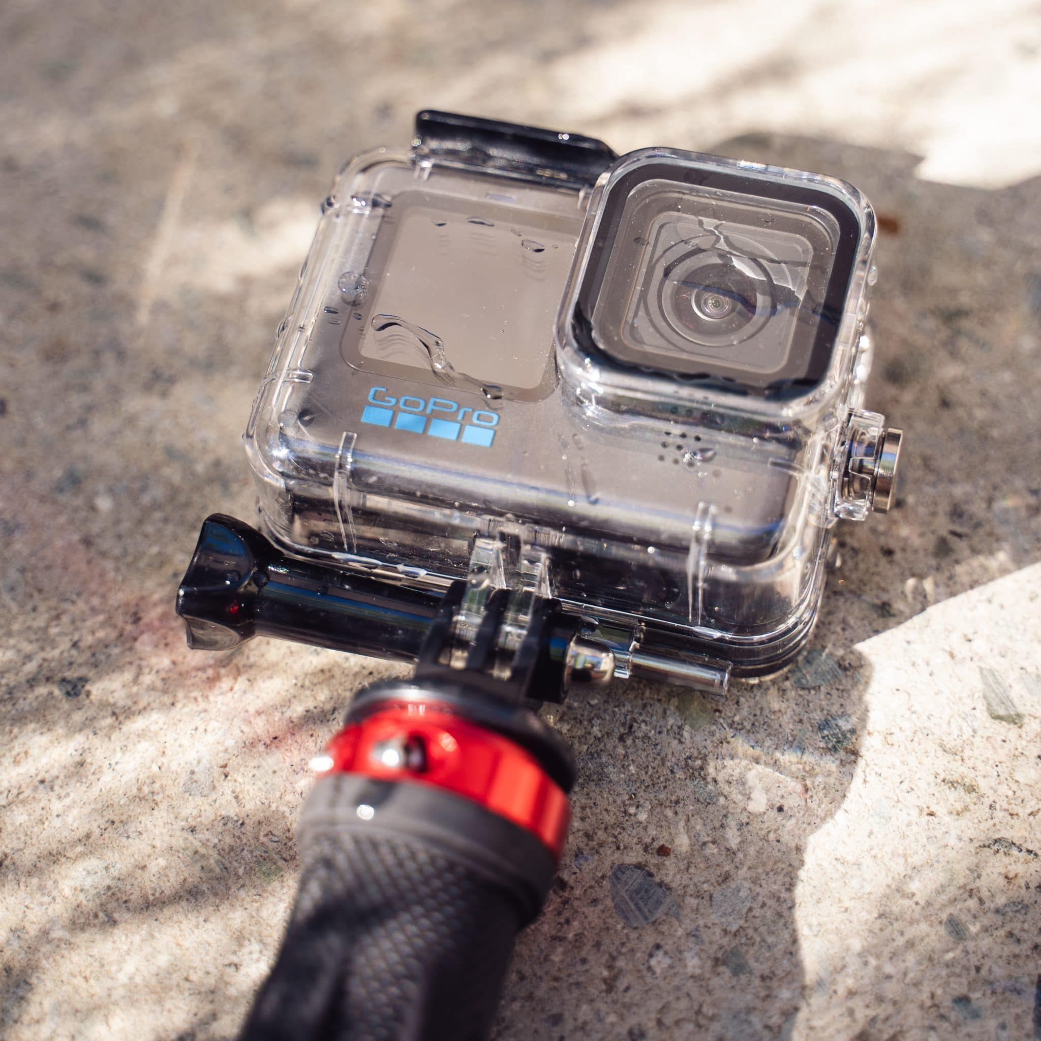 GoPro camera in a waterproof housing, attached to a hand mount, on a stony background.