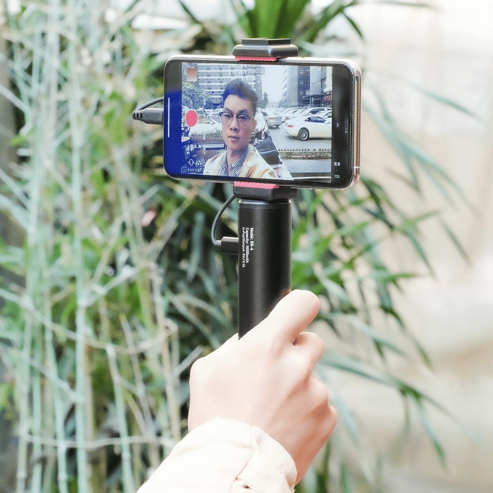 Une main tient une perche à selfie noire avec un smartphone affichant un homme, devant des plantes vertes.