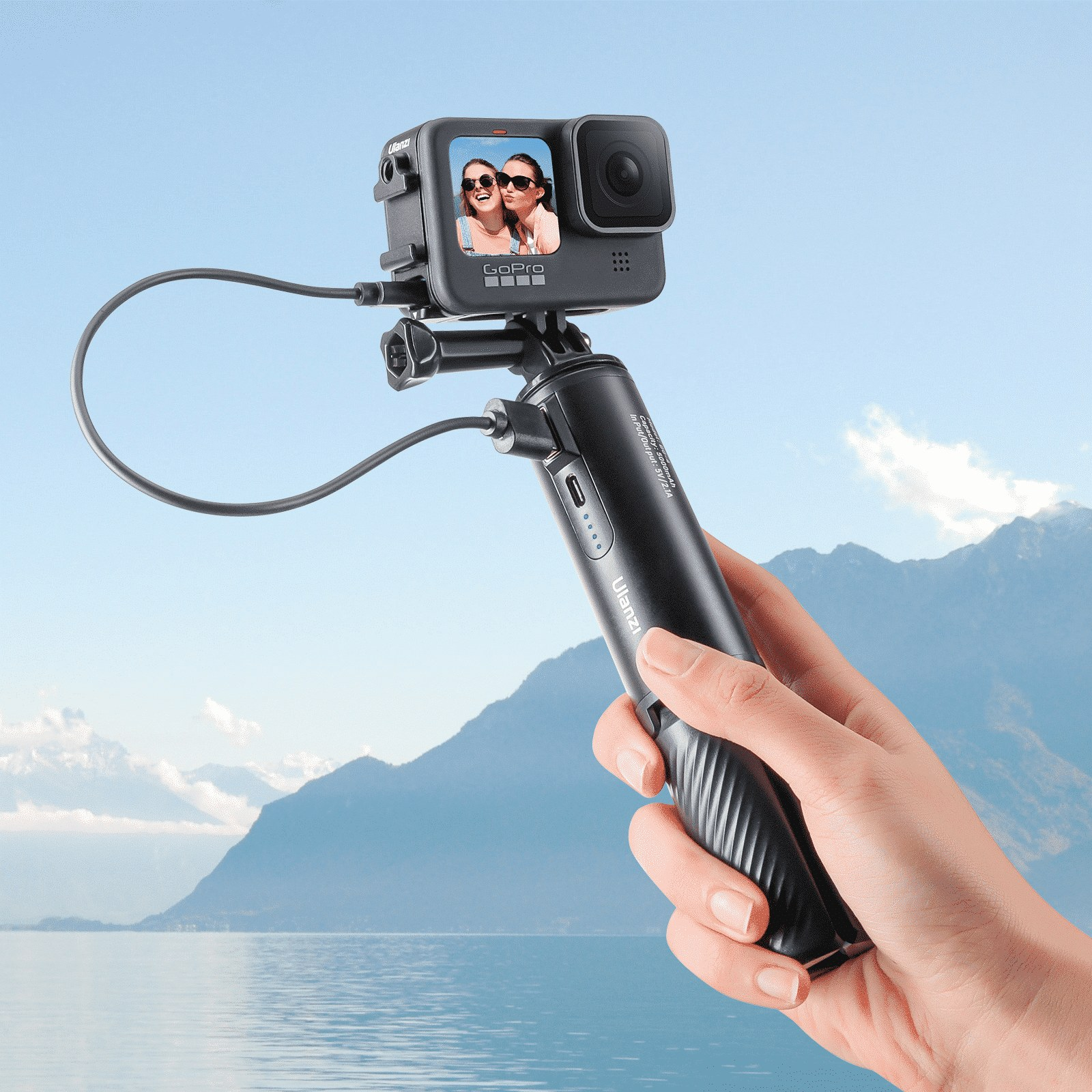 Une main tient une perche à selfie noire avec câble de charge devant un paysage aquatique et montagneux.