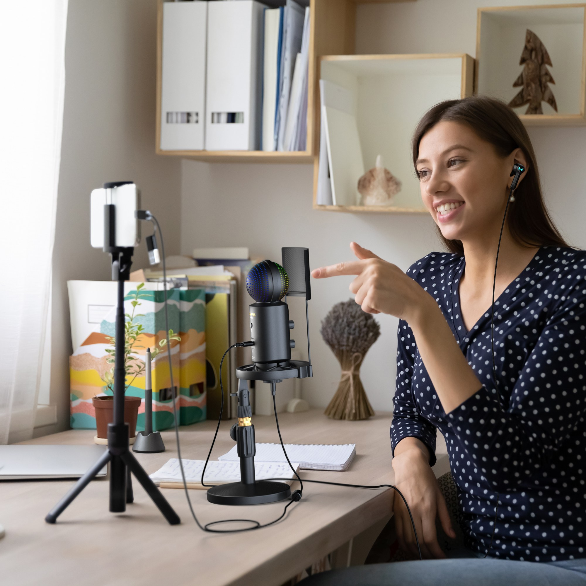 A young woman wearing earphones sits at a desk, pointing at a microphone in front of a smartphone on a tripod.