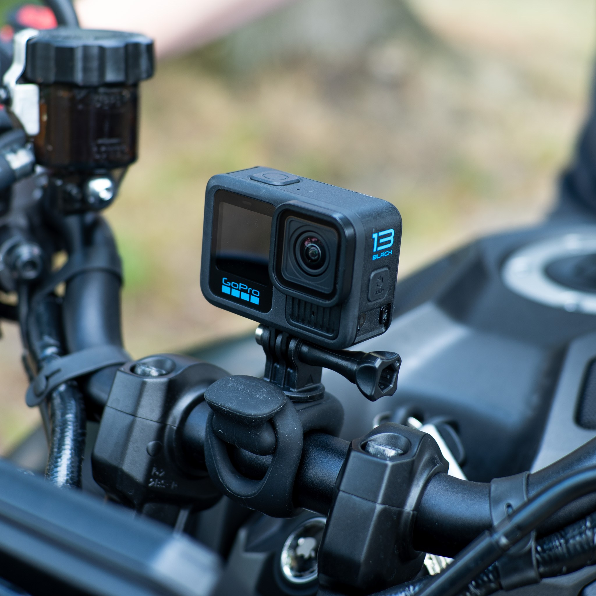 Een GoPro Hero 13 Black actiecamera is gemonteerd op een motorstuur, tegen een wazige natuurlijke achtergrond.