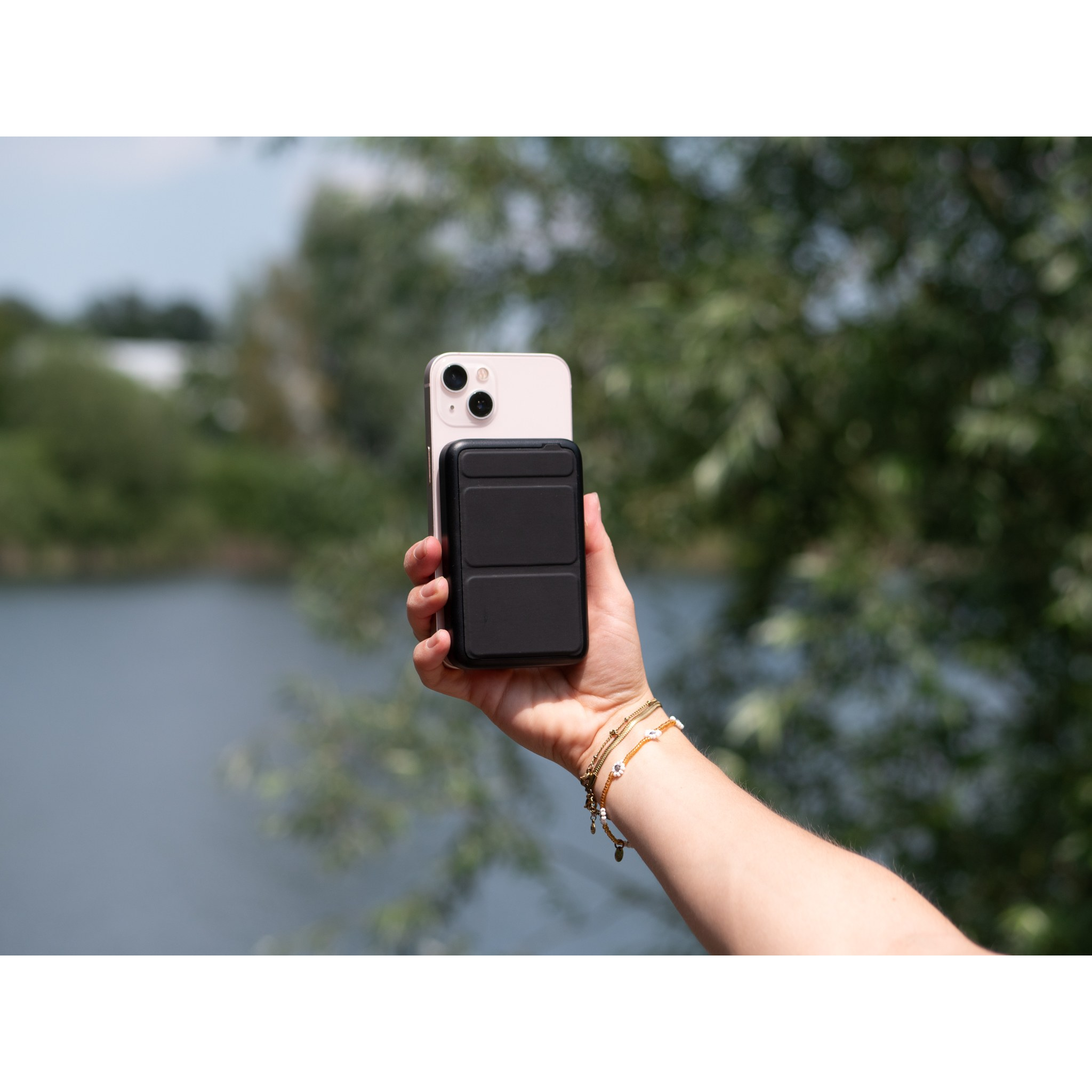 A hand holds a pink smartphone with a black power bank outdoors, with a blurry lake and trees behind.