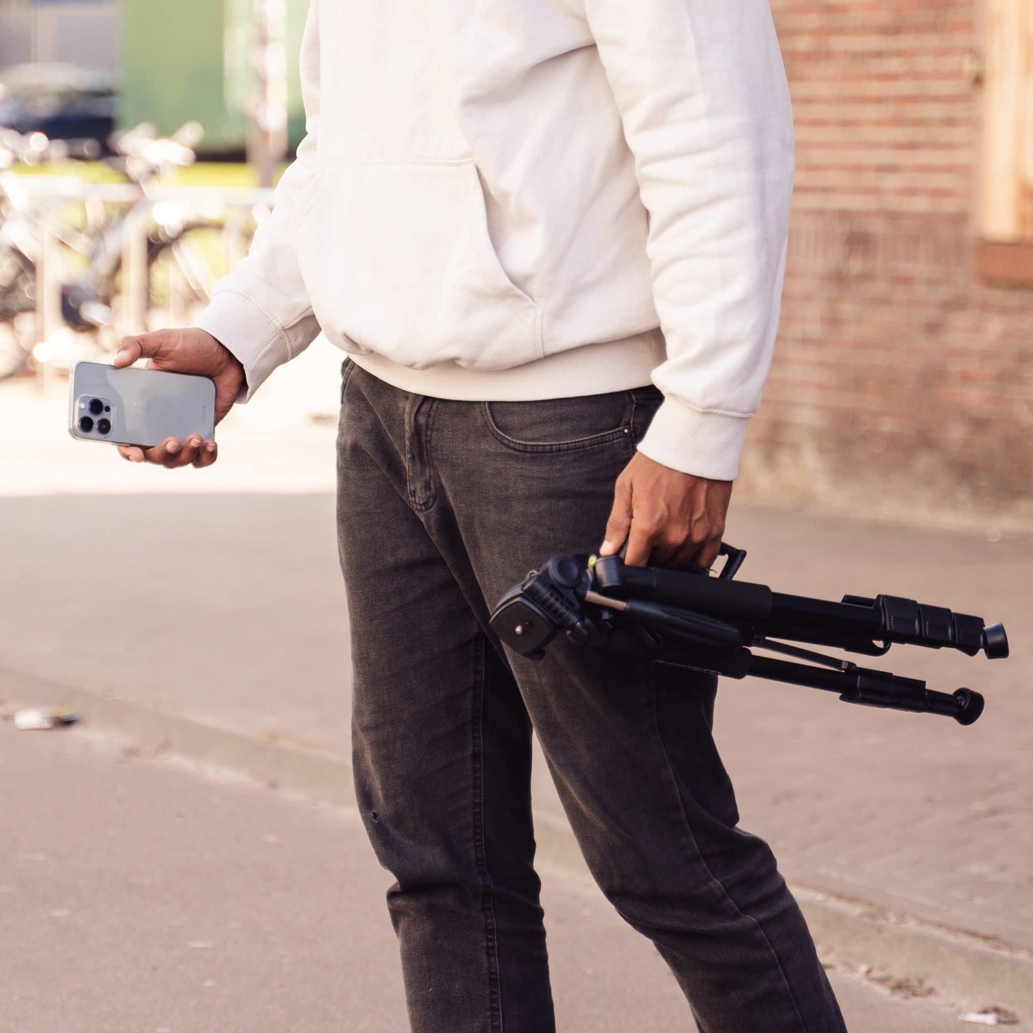 A person in a white hoodie and dark jeans holding a smartphone and a tripod outdoors.