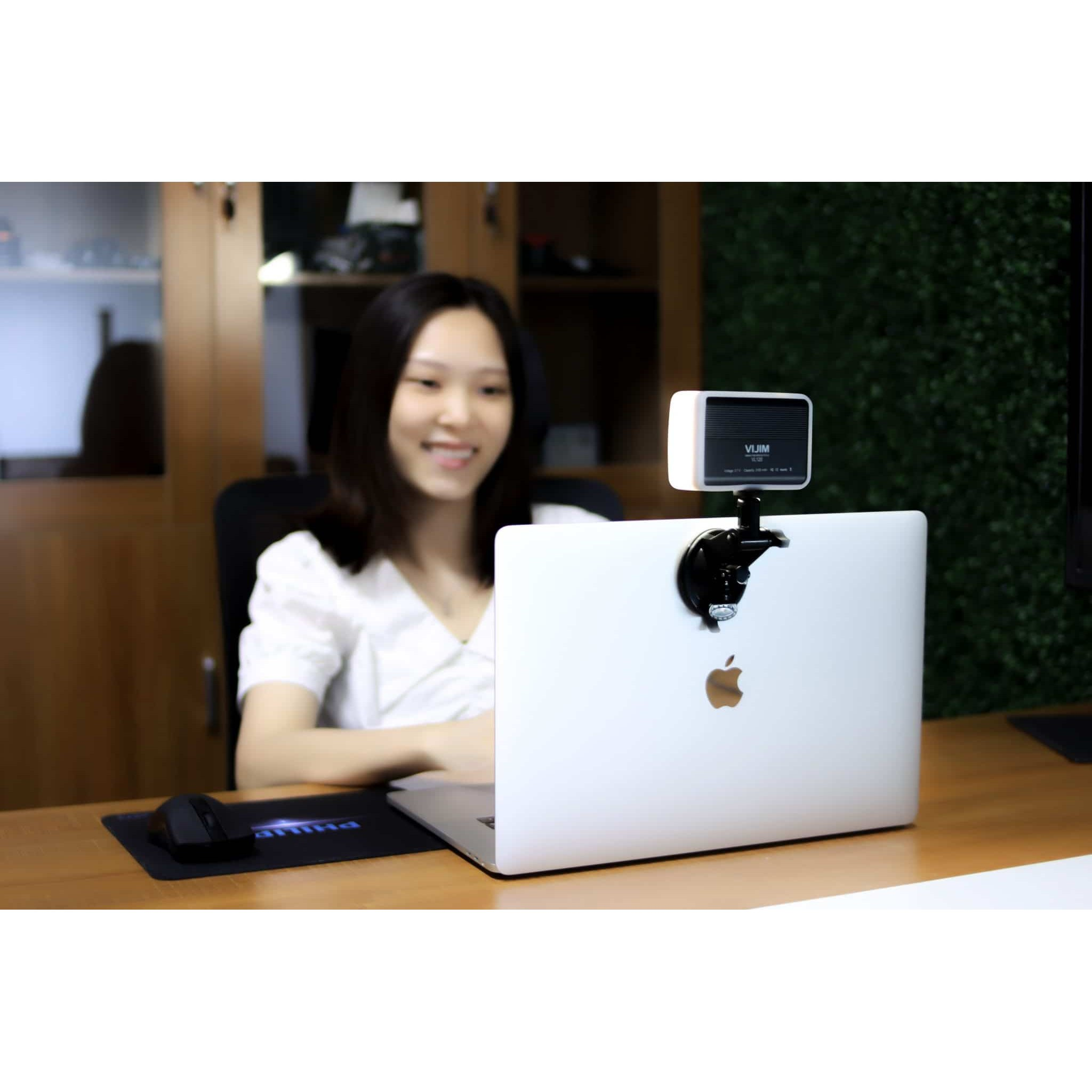 A woman sits at a wooden desk in front of a laptop with an attached LED light.