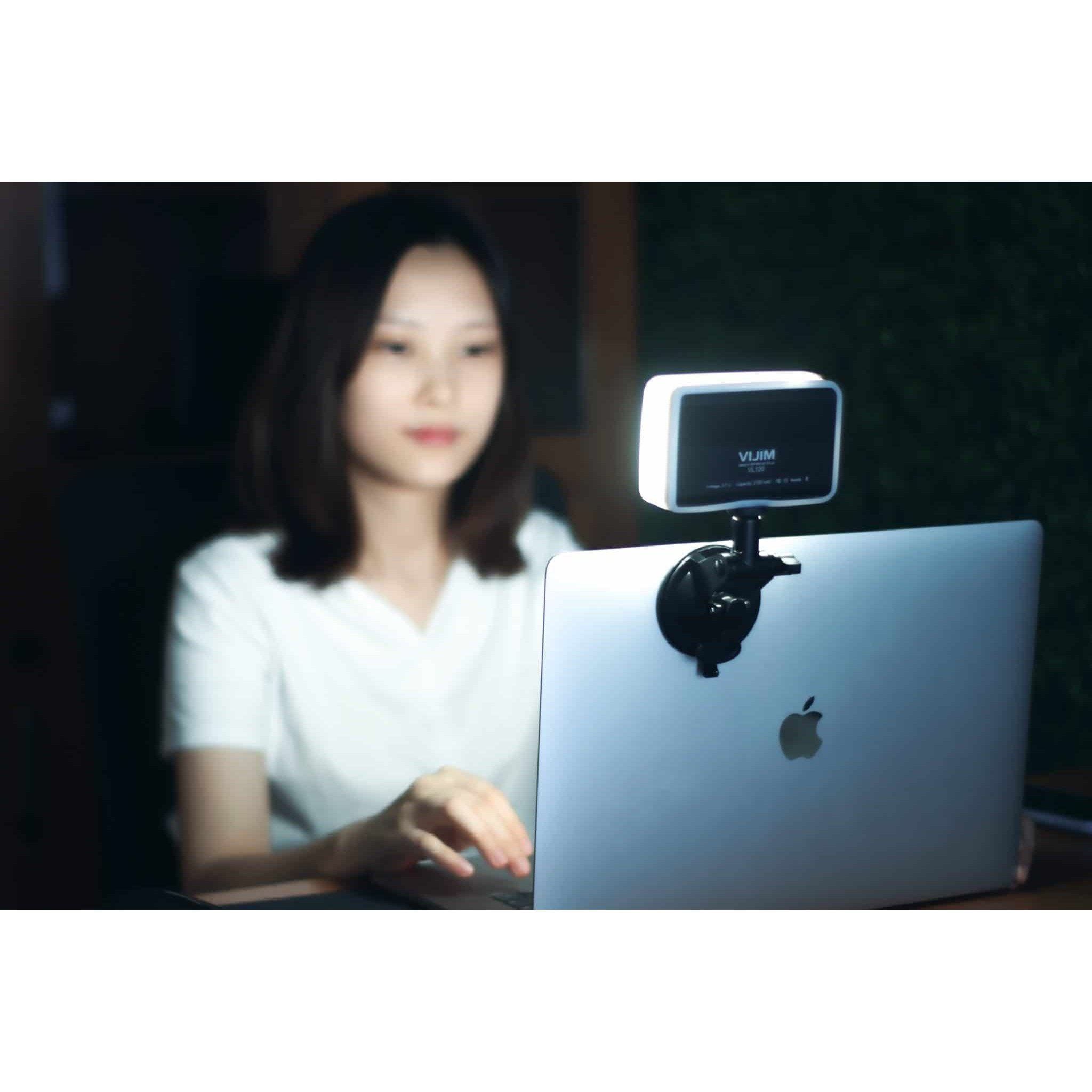 A woman working on a laptop with an attached illuminated LED panel light in a dimly lit room.