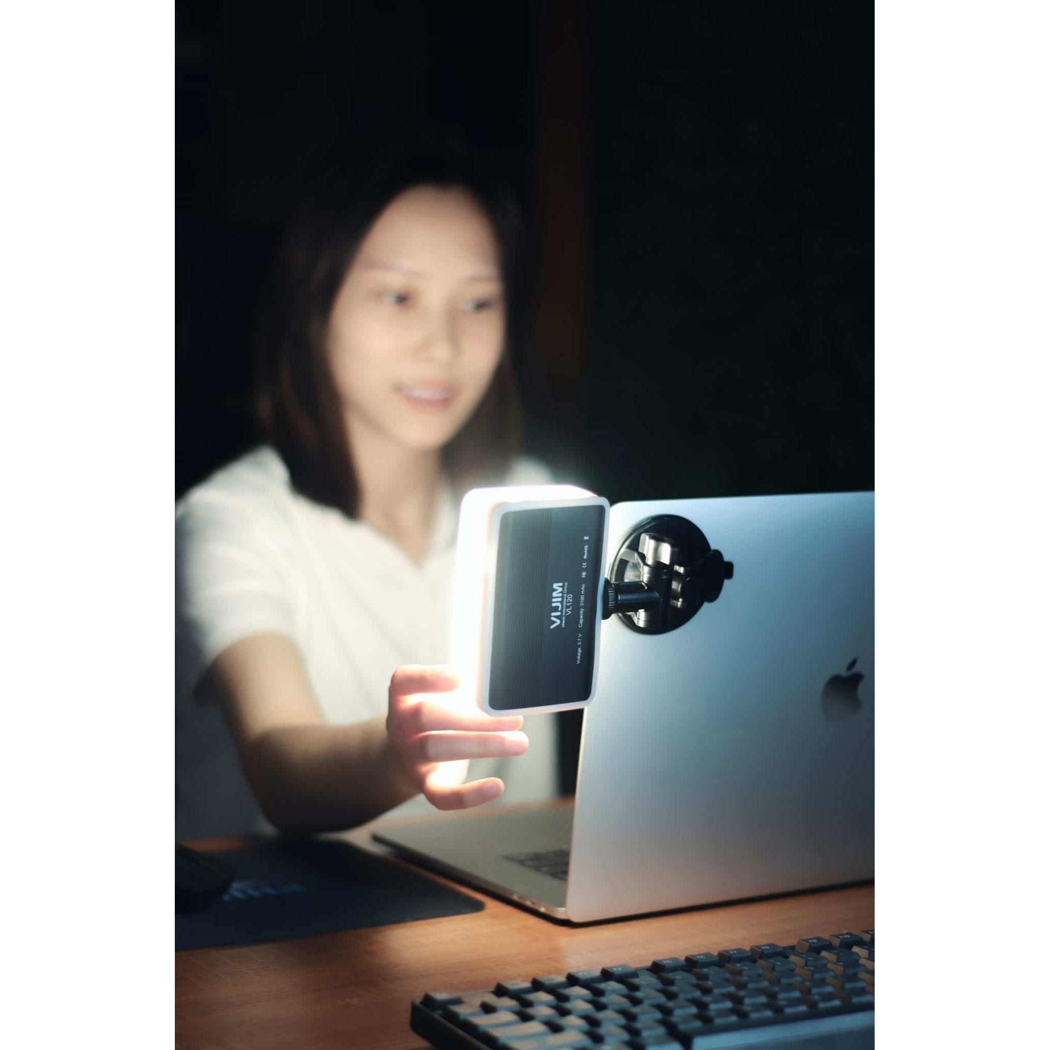 A woman sits at a desk, holding a small LED light in front of a laptop with an attached light mount.