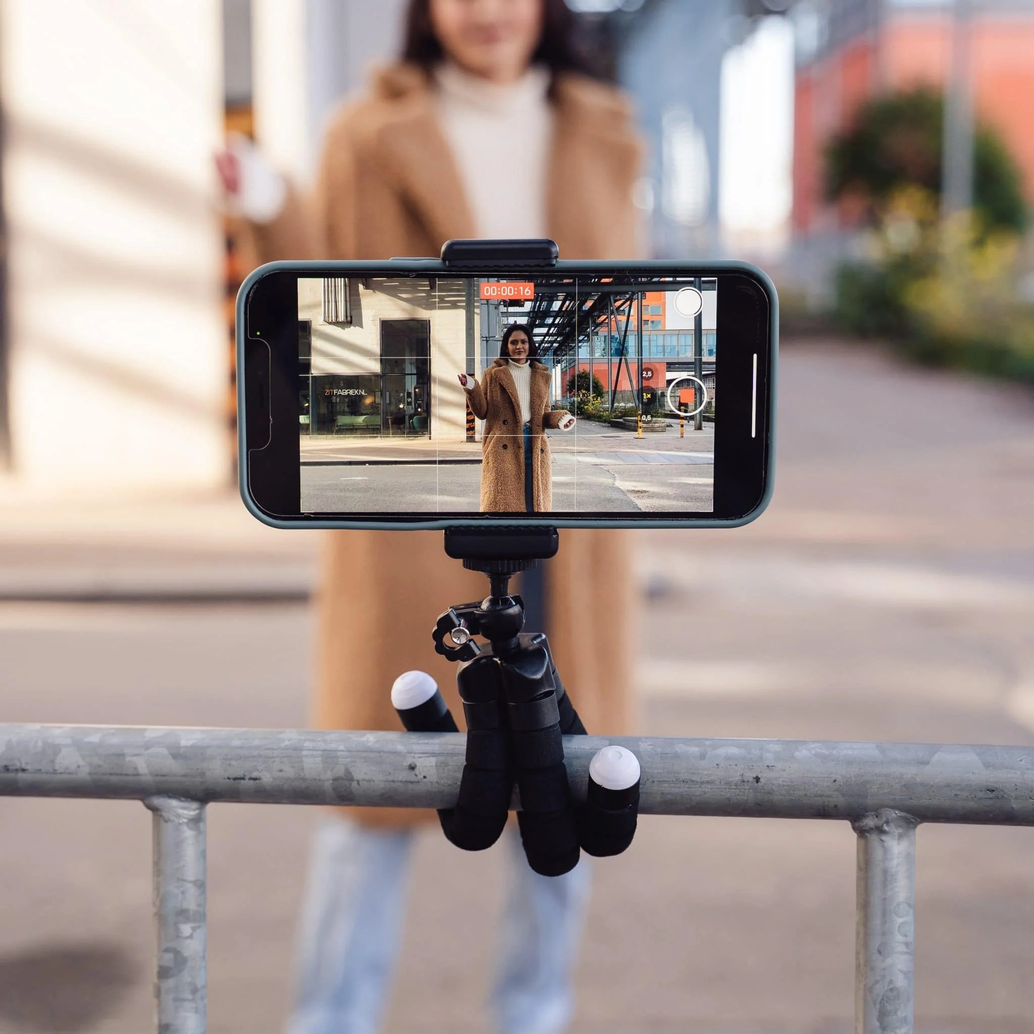 A smartphone on a small tripod attached to a railing records a woman in a coat.