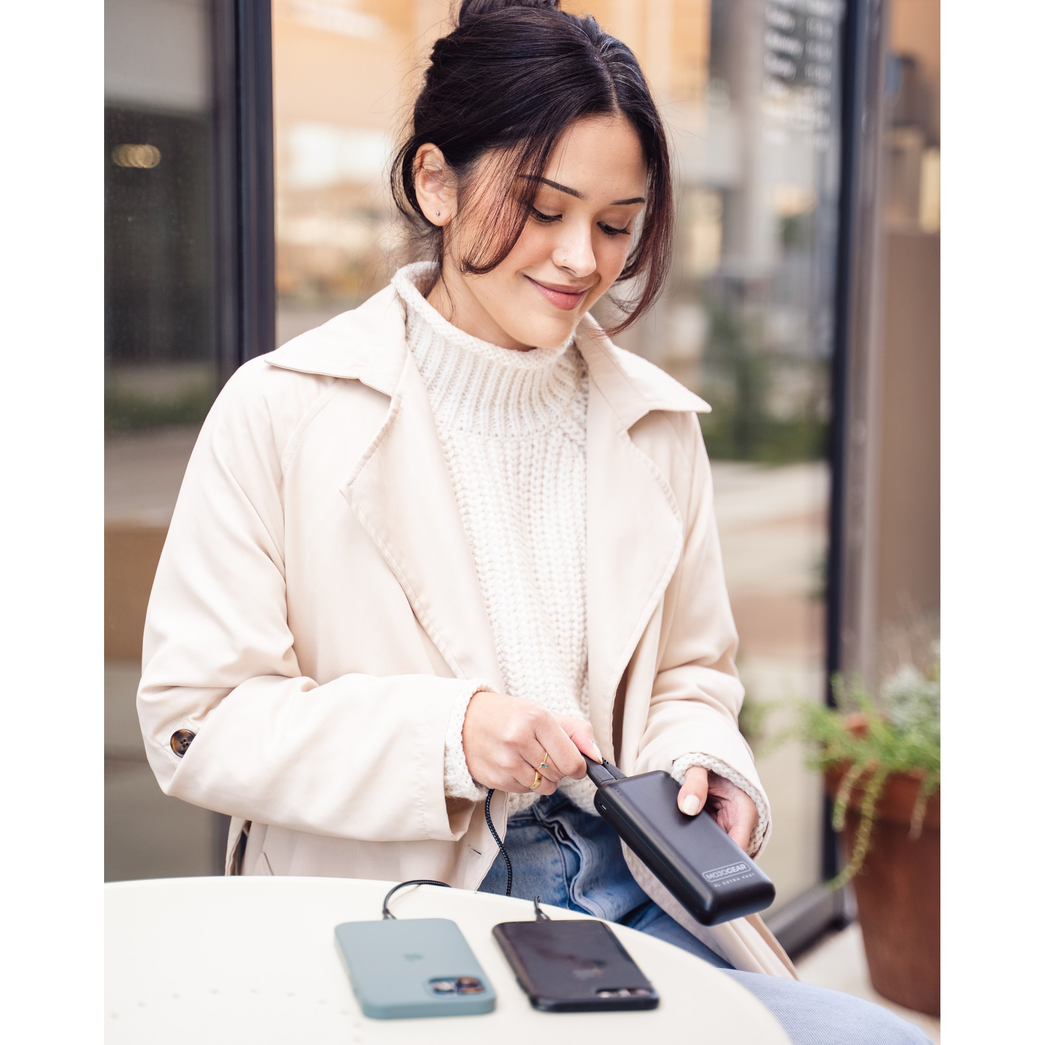 Une femme en manteau beige est assise à une table et connecte une batterie externe noire avec un câble.