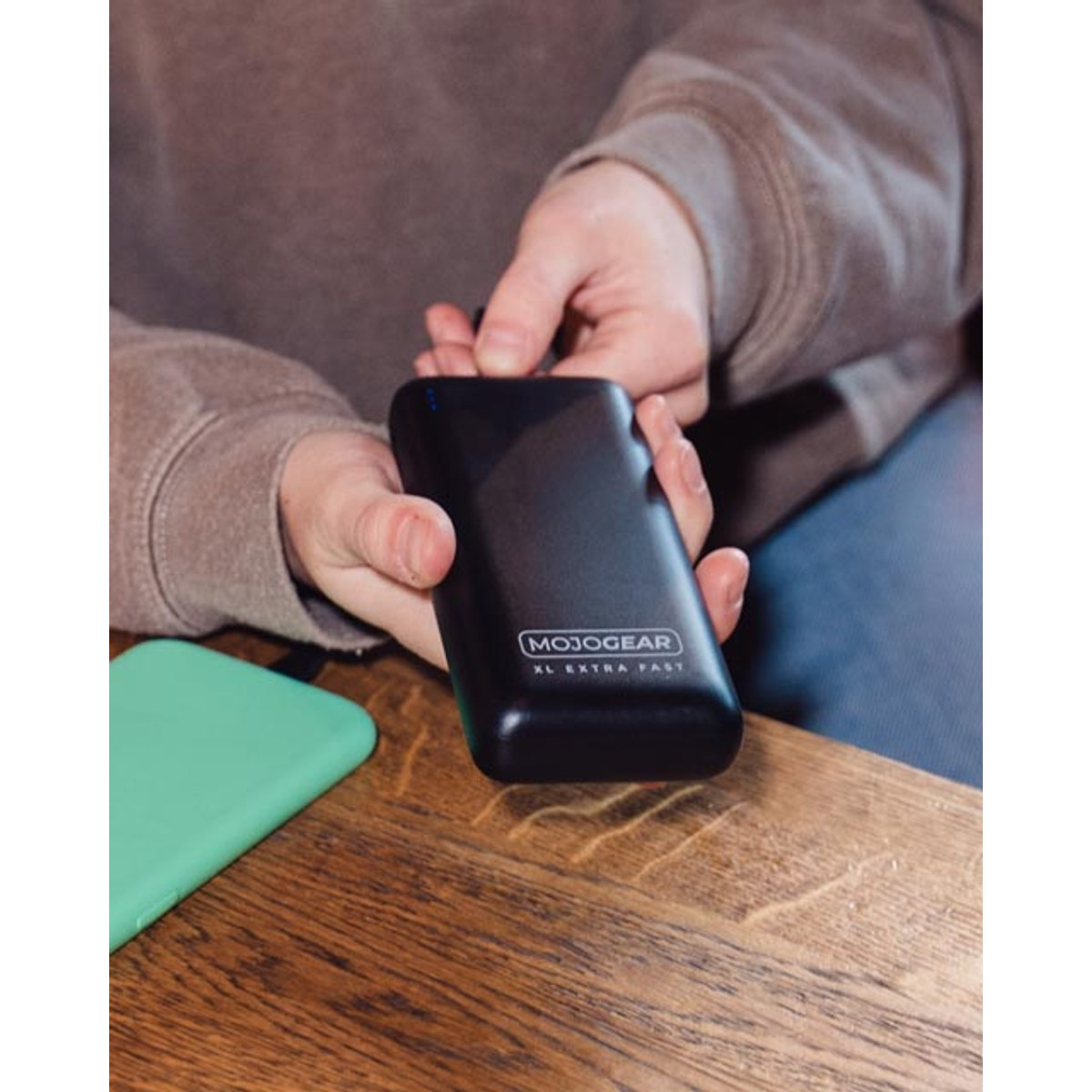 Personne tenant une batterie externe noire Mojo Gear sur une table en bois, avec un smartphone vert à gauche.