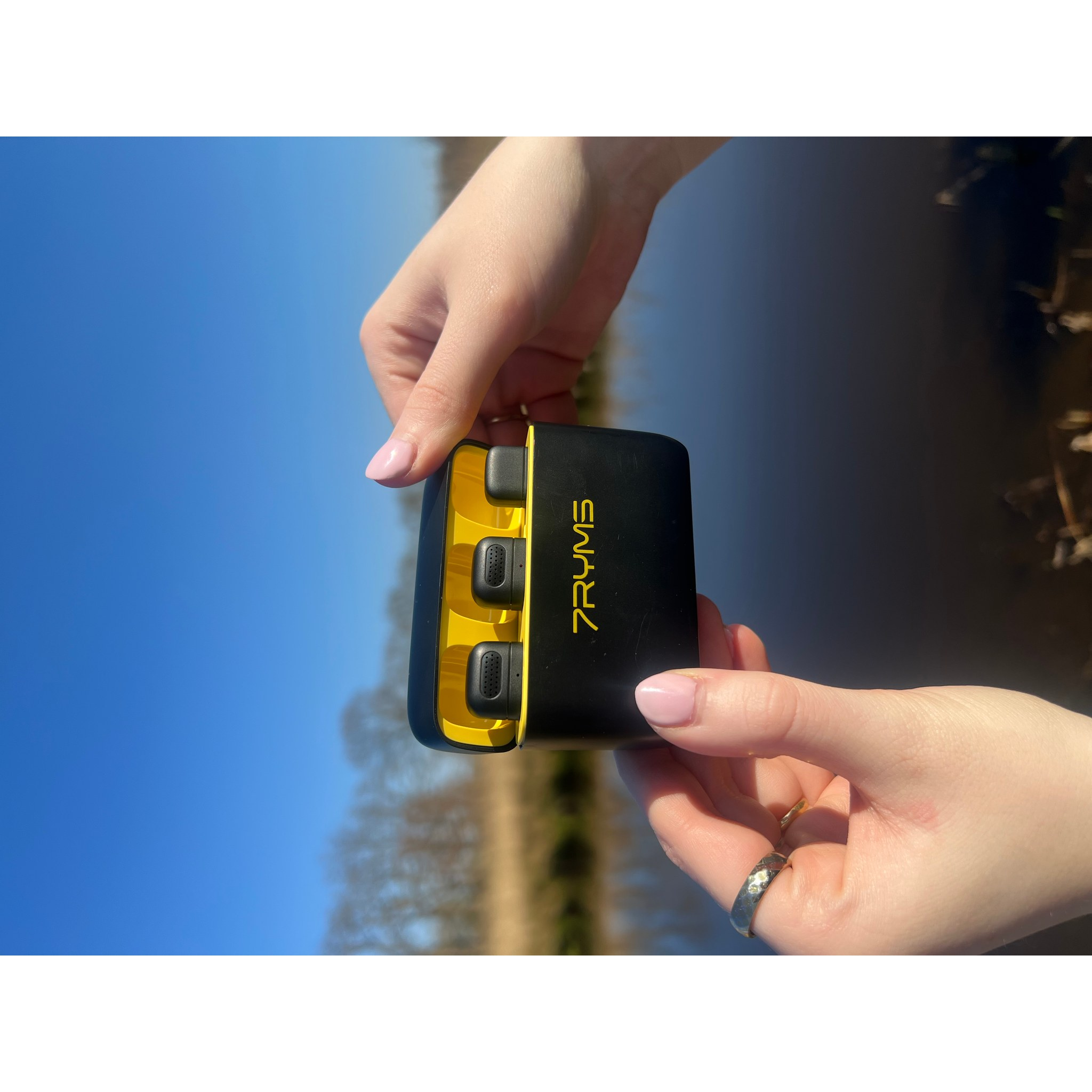 Hände halten ein offenes, schwarz-gelbes Ladecase mit Kopfhörern vor blauem Himmel und Fluss.
