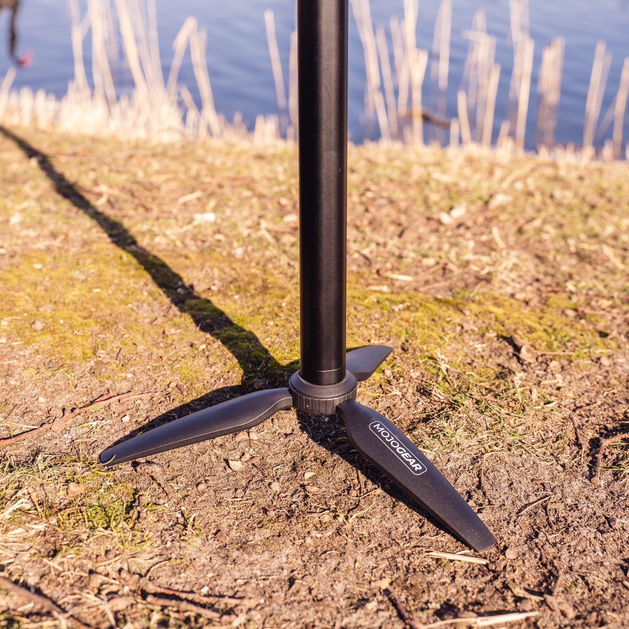 A black MojoGear tripod standing on the ground before a lake.