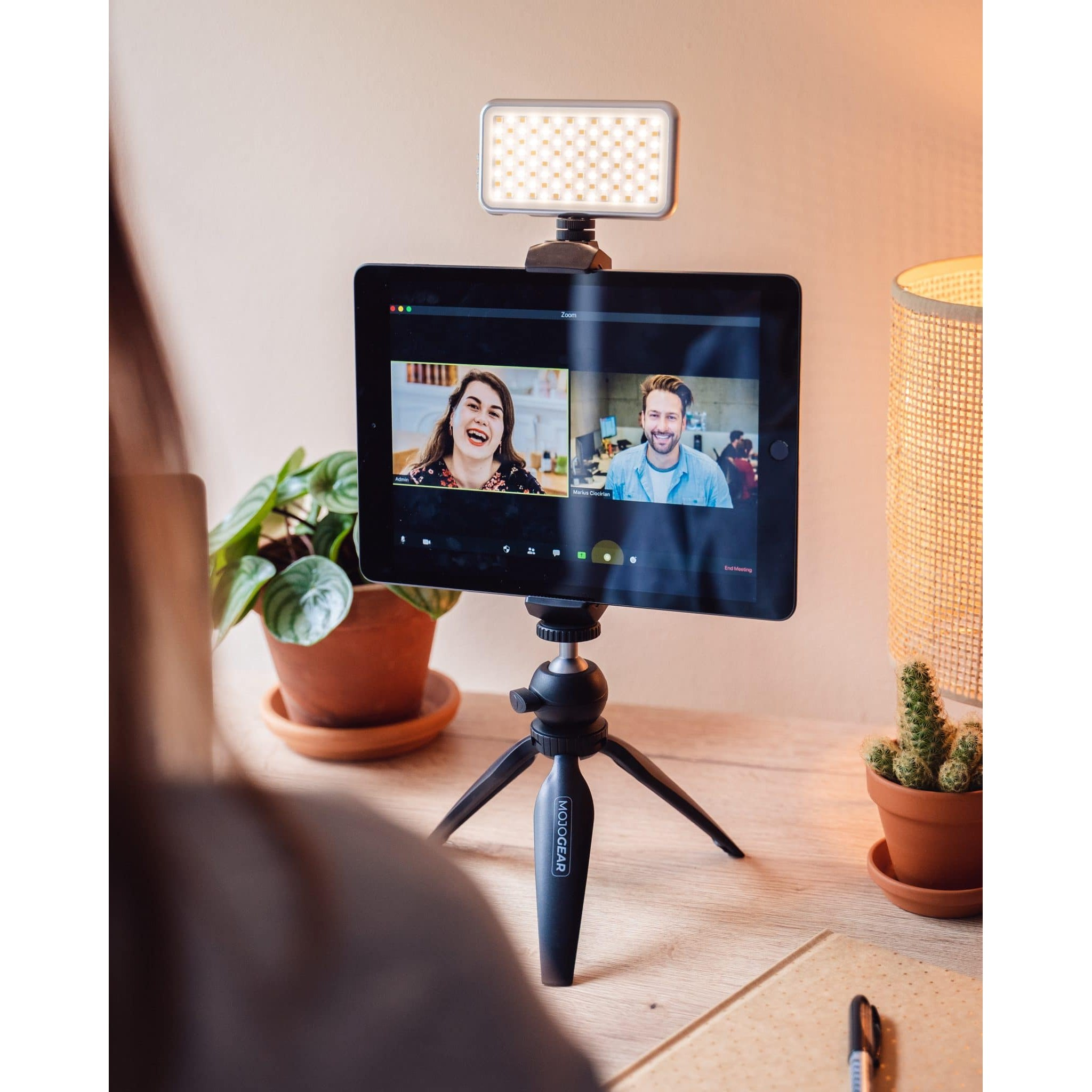Ein Tablet auf einem Minitripod zeigt einen Videoanruf mit zwei Personen, mit Ringleuchte, auf einem Holzschreibtisch.