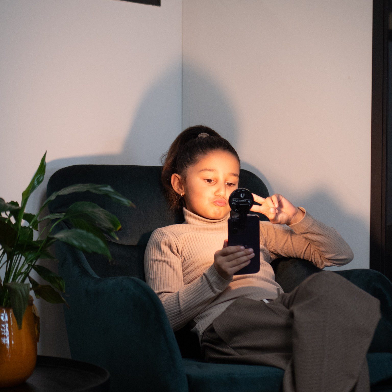 A young girl in a beige sweater sits in a green armchair, holding a phone with a light and flashing a peace sign.