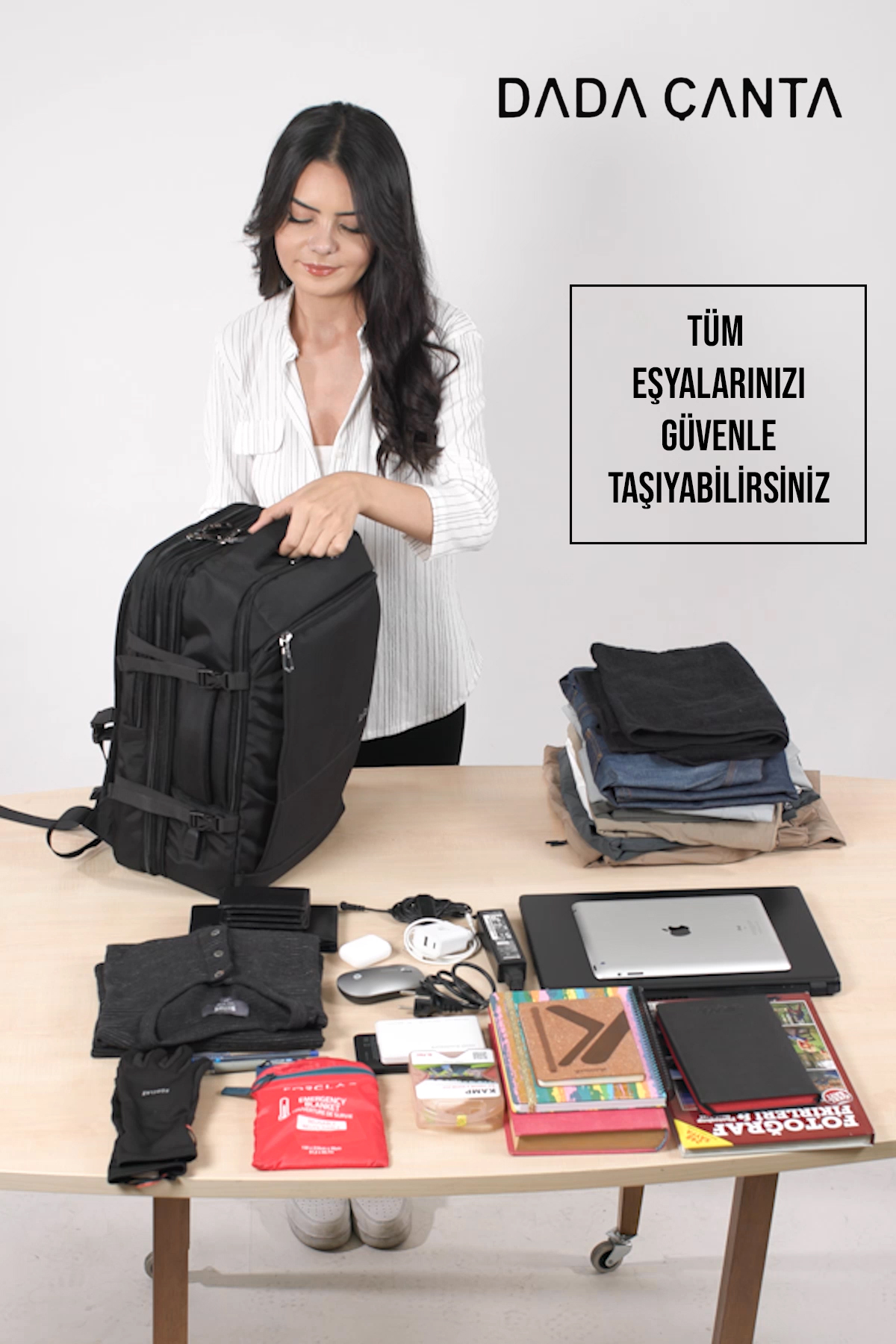 Bir kadın, giysiler, elektronik eşyalar, kitaplar ve aksesuarlarla dolu masada siyah büyük bir sırt çantası topluyor.