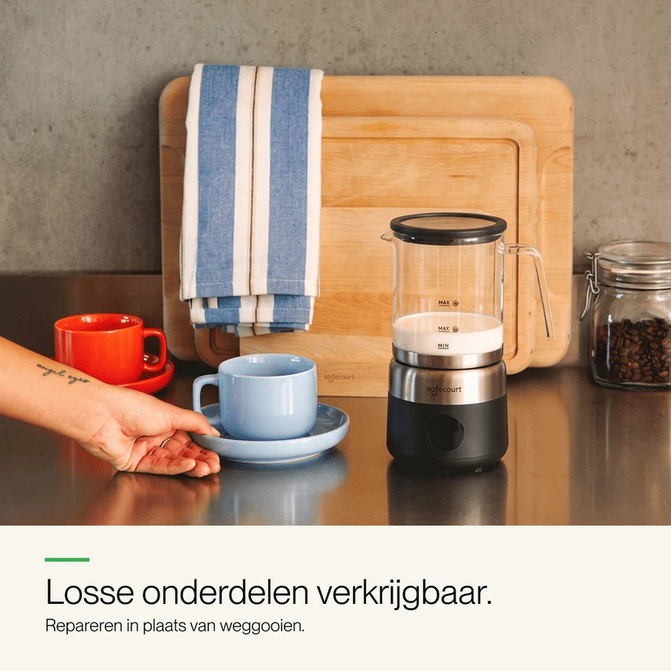 Een melkopschuimer, blauwe en rode kopjes op een aanrecht, met houten snijplanken en een gestreepte handdoek.