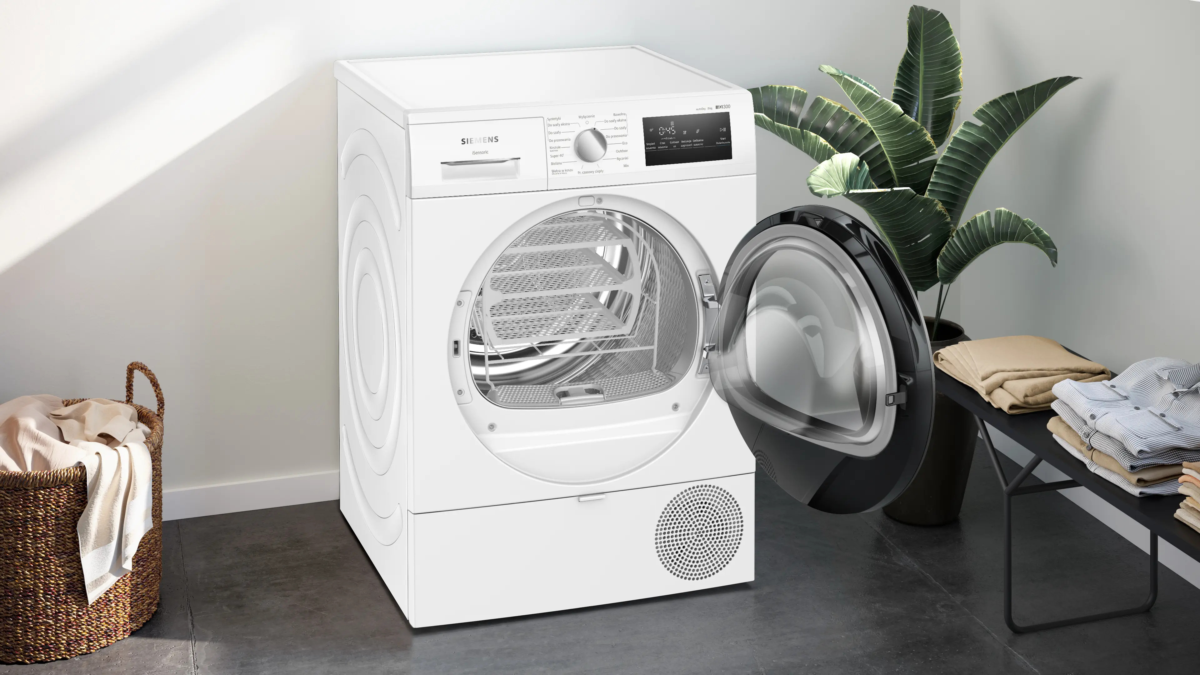 Biała suszarka Siemens z otwartymi czarnymi drzwiami i koszem w środku. Po prawej roślina i ławka z praniem.