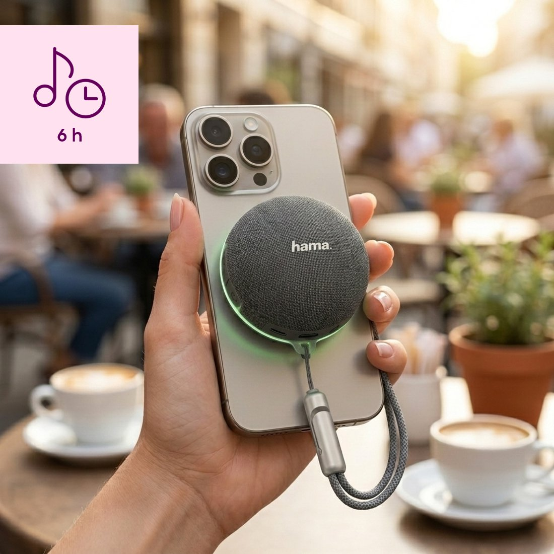 Ręka trzyma smartfon z magnetycznym, szarym głośnikiem Bluetooth Hama i zintegrowaną smyczą w kawiarni.
