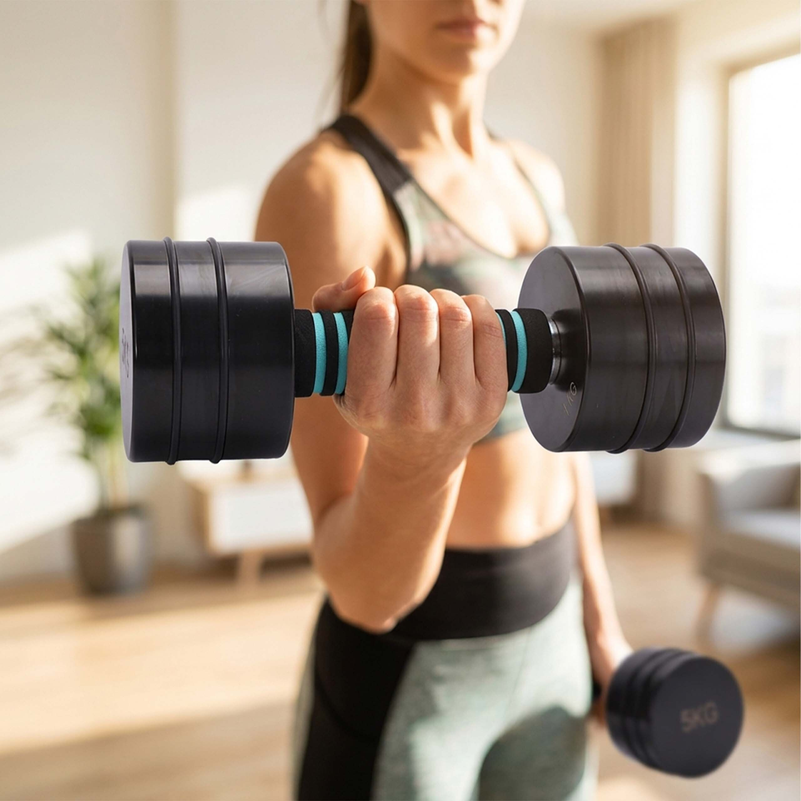 Eine Frau in Sportkleidung hält in jedem Arm eine schwarze Fünf-Kilo-Hantel in einem hellen Raum.