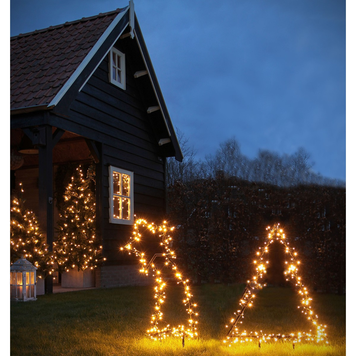 Eine dunkle Holzhütte im Garten ist abends mit warmen Lichterketten und leuchtenden Figuren geschmückt.