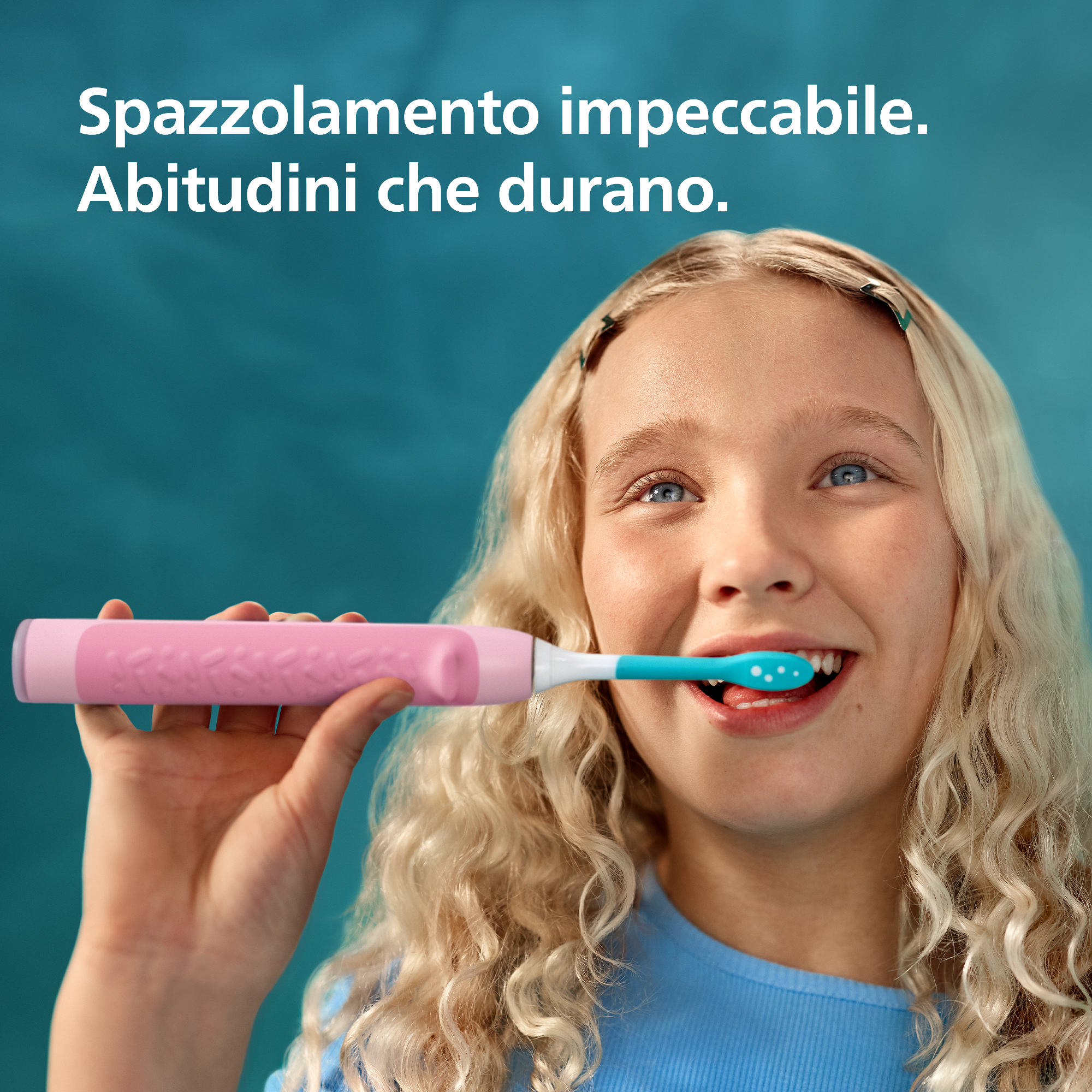 Una bambina bionda sorride e si lava i denti con uno spazzolino elettrico rosa su sfondo blu.