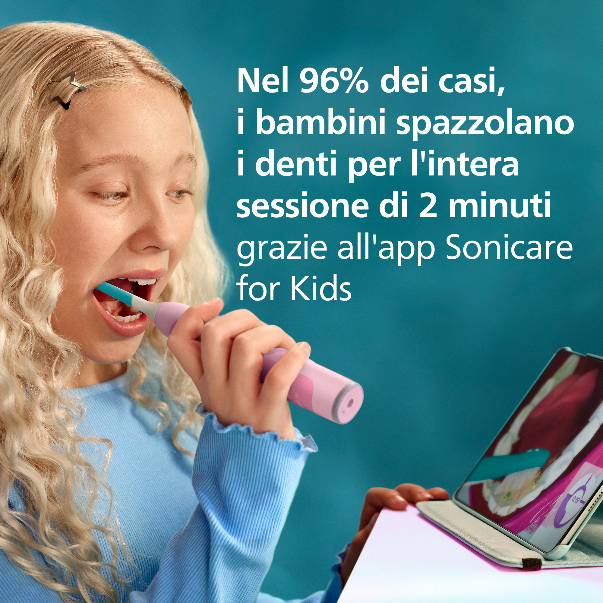 Una bambina bionda in blu si lava i denti con uno spazzolino elettrico rosa guardando un tablet.