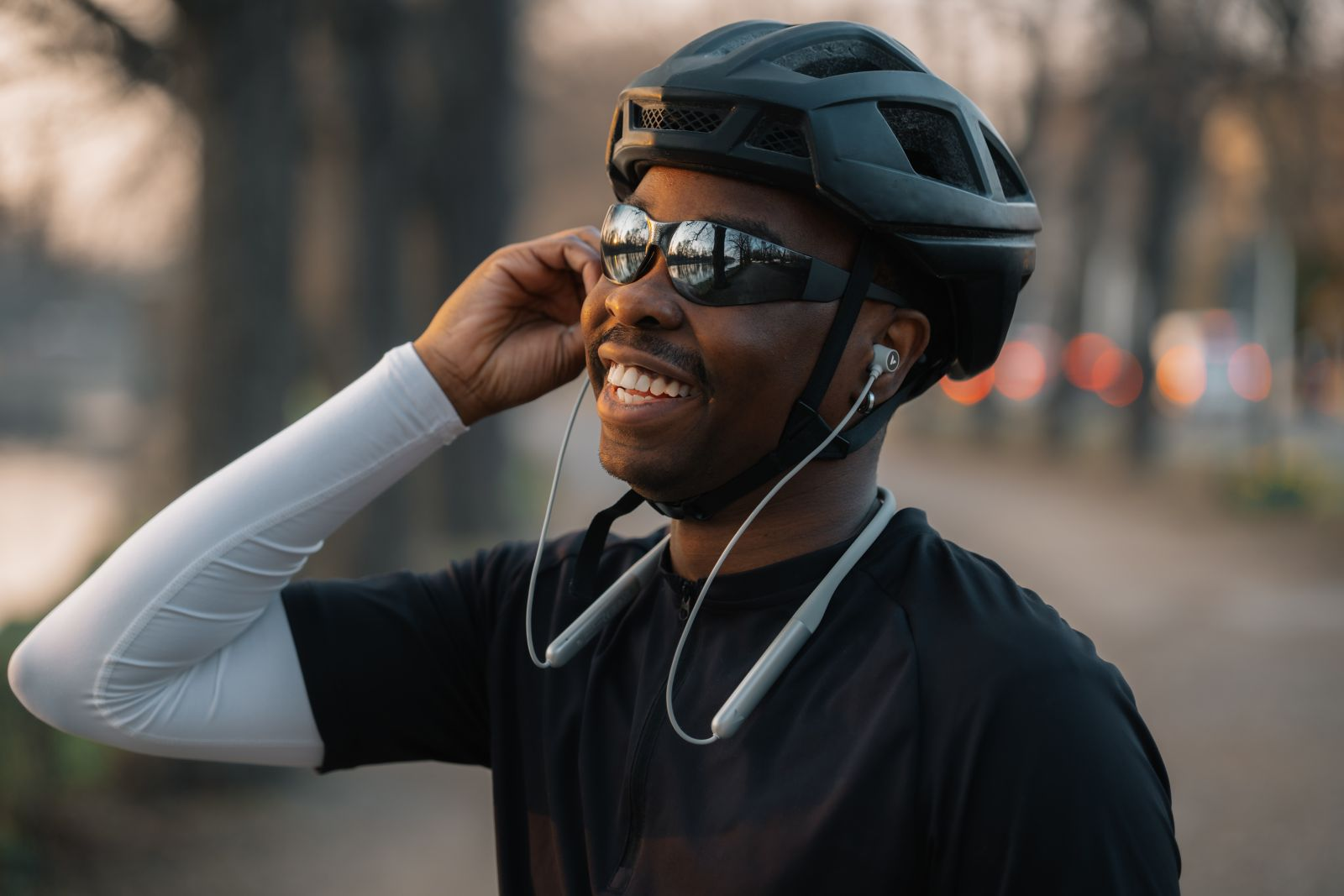 Un ciclista sonriente con casco negro, gafas de sol y auriculares de banda para el cuello.