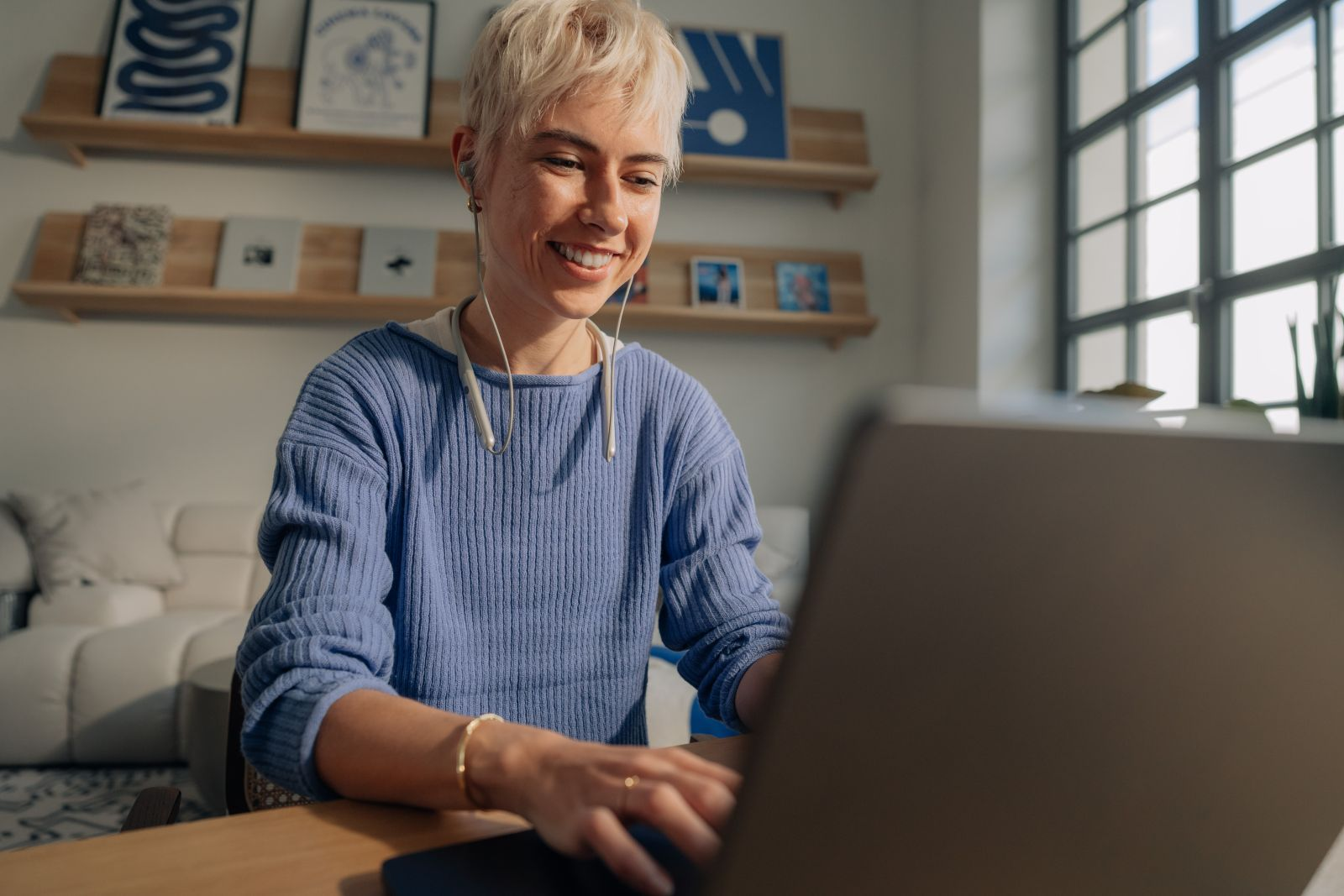 Una mujer rubia con suéter azul escribe en una laptop en una habitación luminosa.