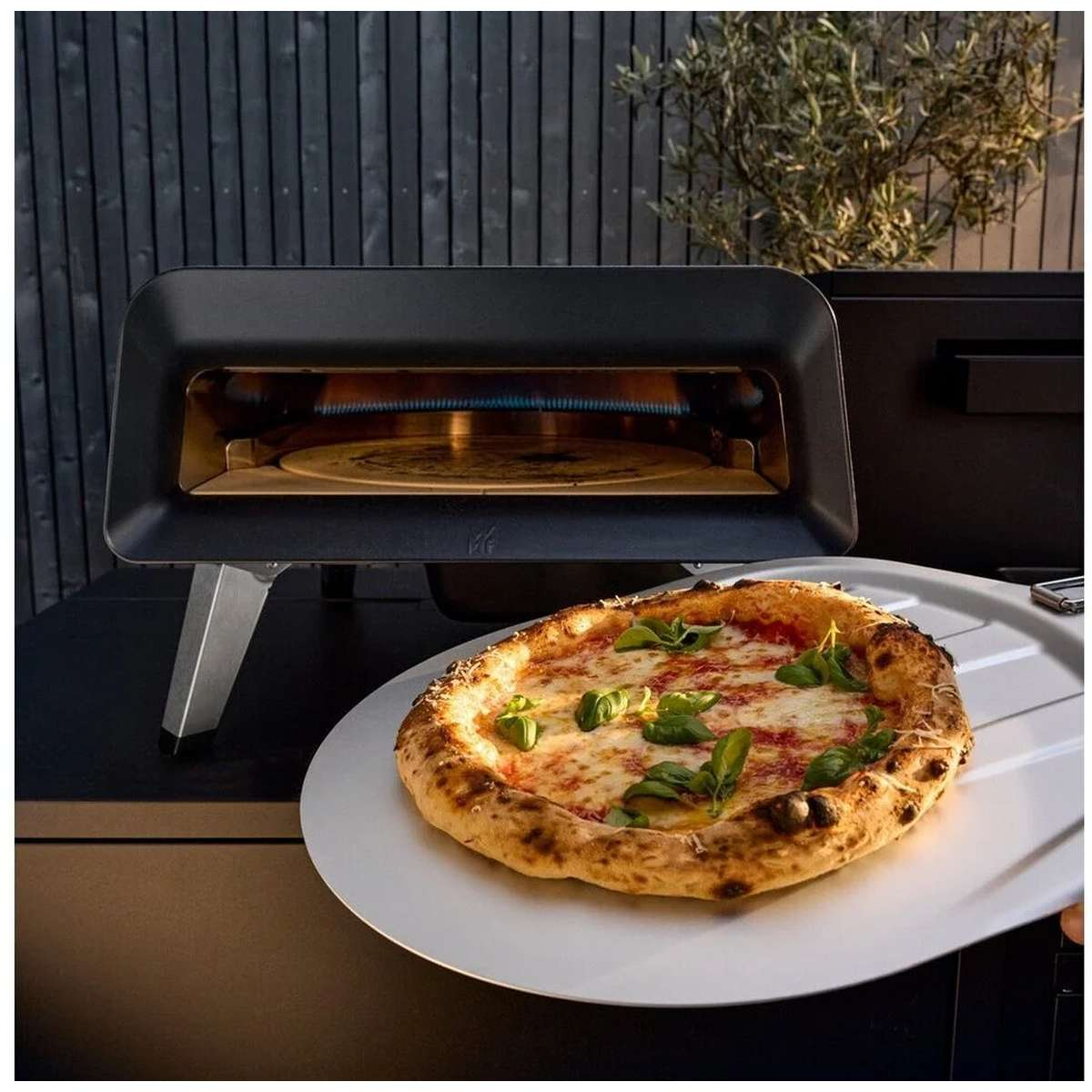 Pizza appena sfornata con formaggio fuso e foglie di basilico su un piatto davanti a un forno per pizza.