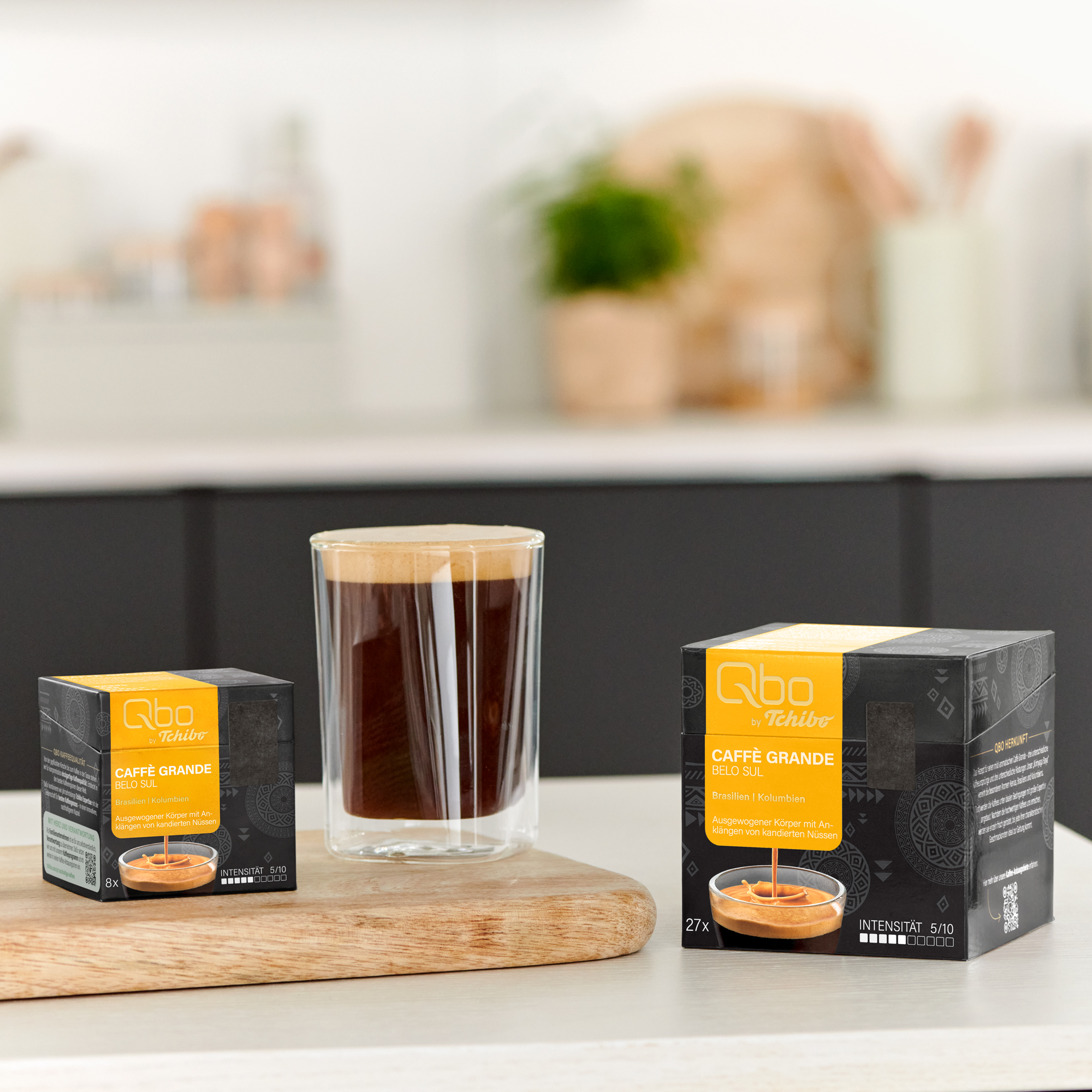 Glas Kaffee und Kaffeekapseln auf einem Holzbrett vor einer Küche.
