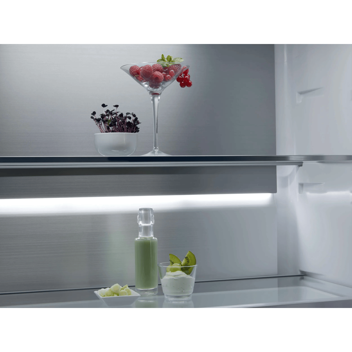 Dentro de un frigorífico iluminado, estantes de cristal sostienen una copa con bayas, una planta, zumo y yogur.