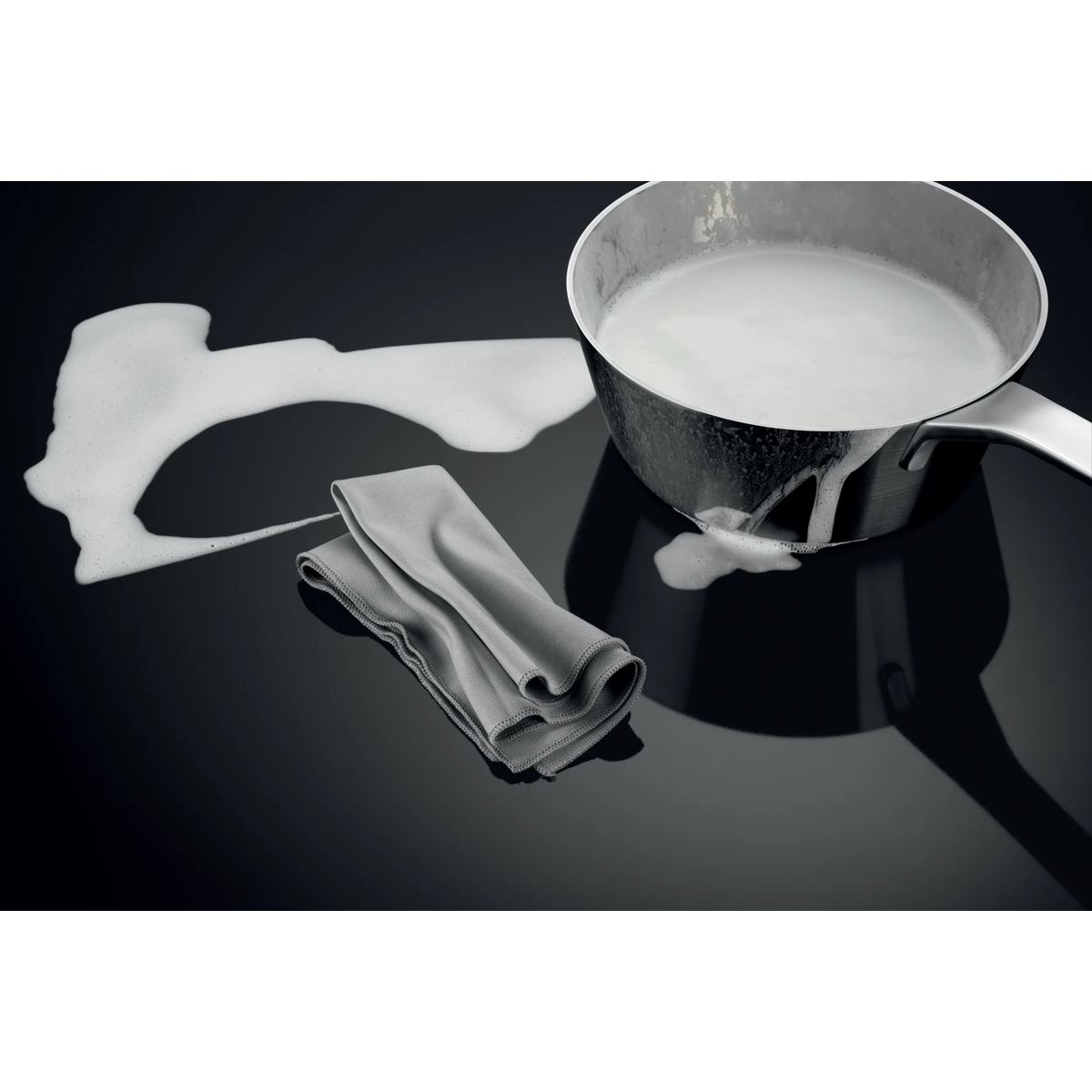 Una pentola di metallo con latte bollente, un panno grigio e schiuma su un piano cottura.