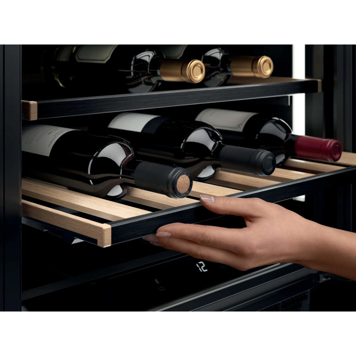 Una mano estrae un ripiano di legno in una cantinetta per vini piena di bottiglie.