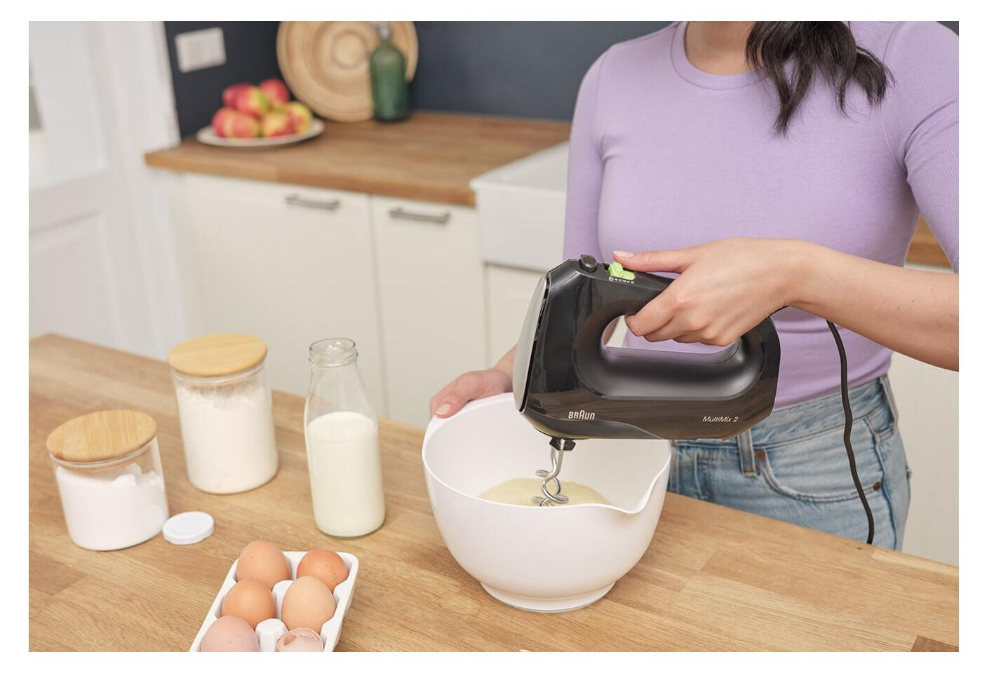 A woman in a purple top mixes batter using a black hand mixer in a white bowl on a wooden counter.
