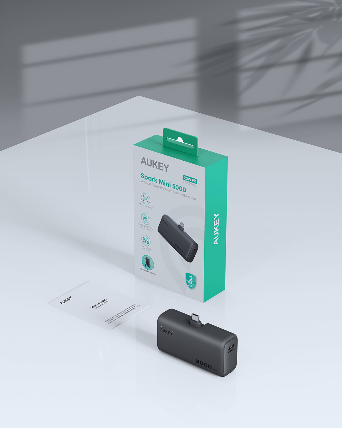 Beyaz bir masada, turkuaz kutusu ve kullanım kılavuzunun yanında duran bir Aukey Spark Mini 5000 taşınabilir şarj cihazı.